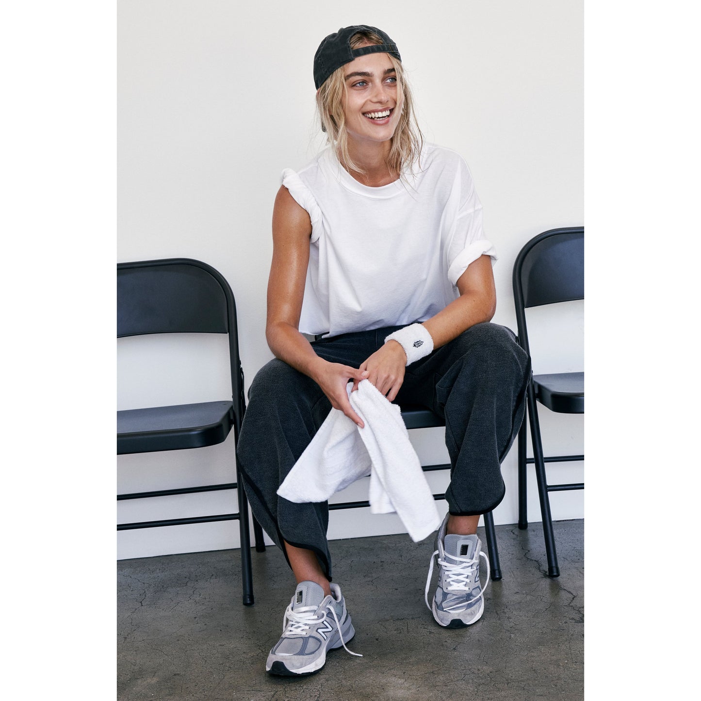 Woman in casual attire smiling, sitting on a bench with a towel on her lap, wearing an oversized boxy fit white Free People Movement Inspire Tee, black cap, and grey sneakers.