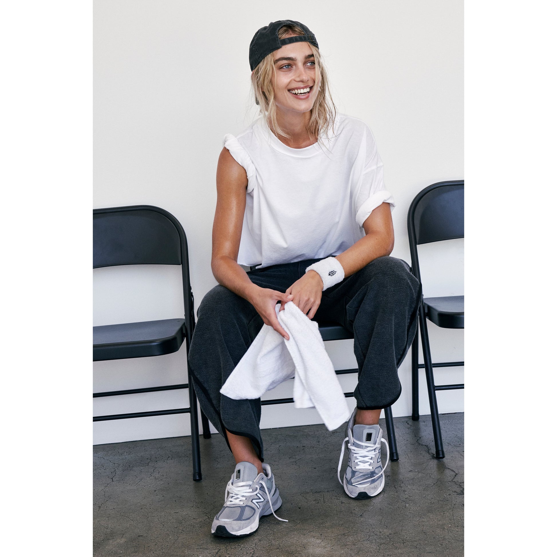 Woman in casual attire smiling, sitting on a bench with a towel on her lap, wearing an oversized boxy fit white Free People Movement Inspire Tee, black cap, and grey sneakers.