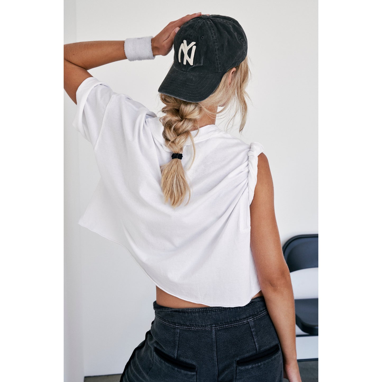 Woman from behind wearing a black NY cap and white oversized boxy fit Free People Movement Inspire Tee knotted at the waist, having a ponytail. Black jeans visible.