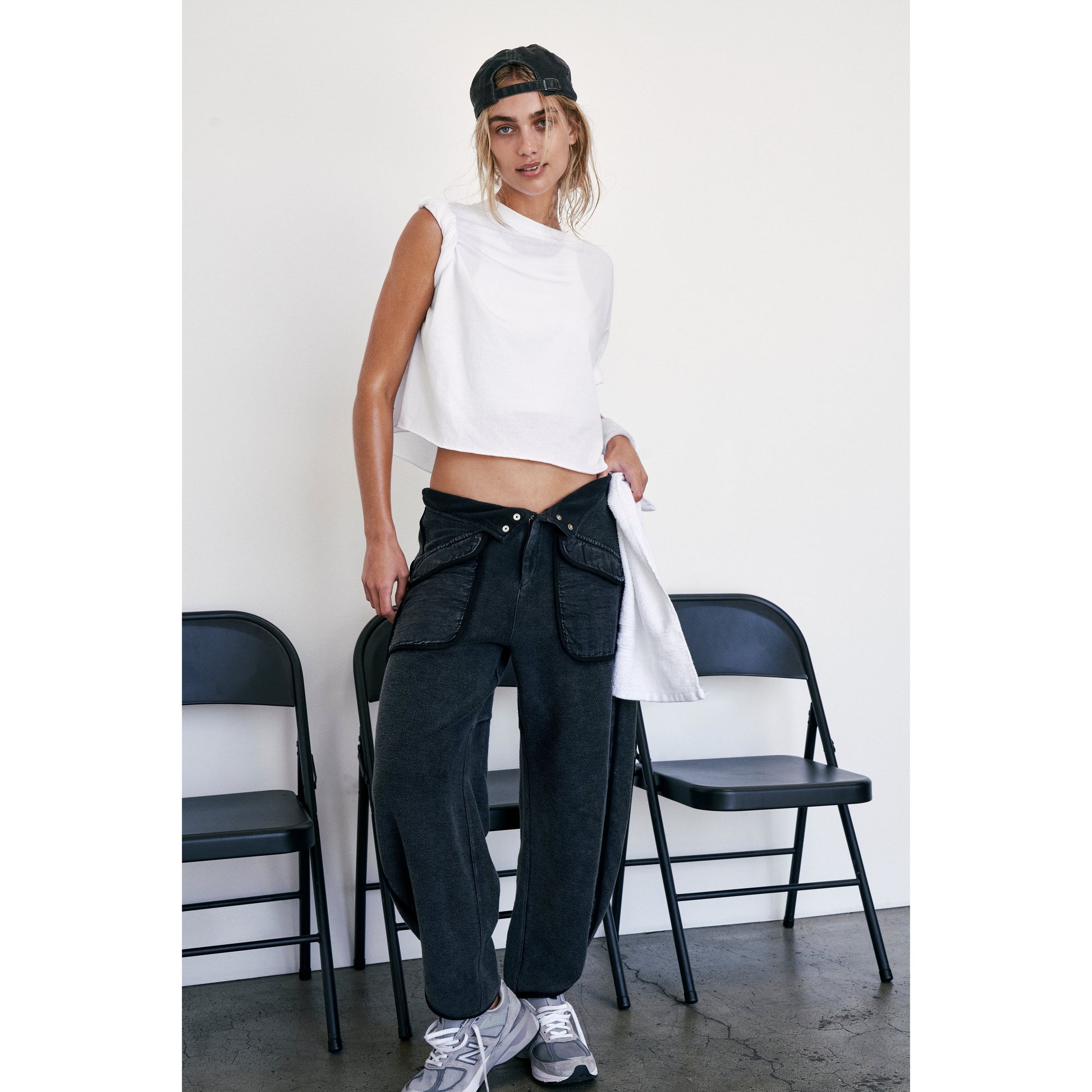 Young woman in a Free People Movement white Inspire Tee and dark jeans standing beside metal chairs, wearing a backward cap and sneakers.