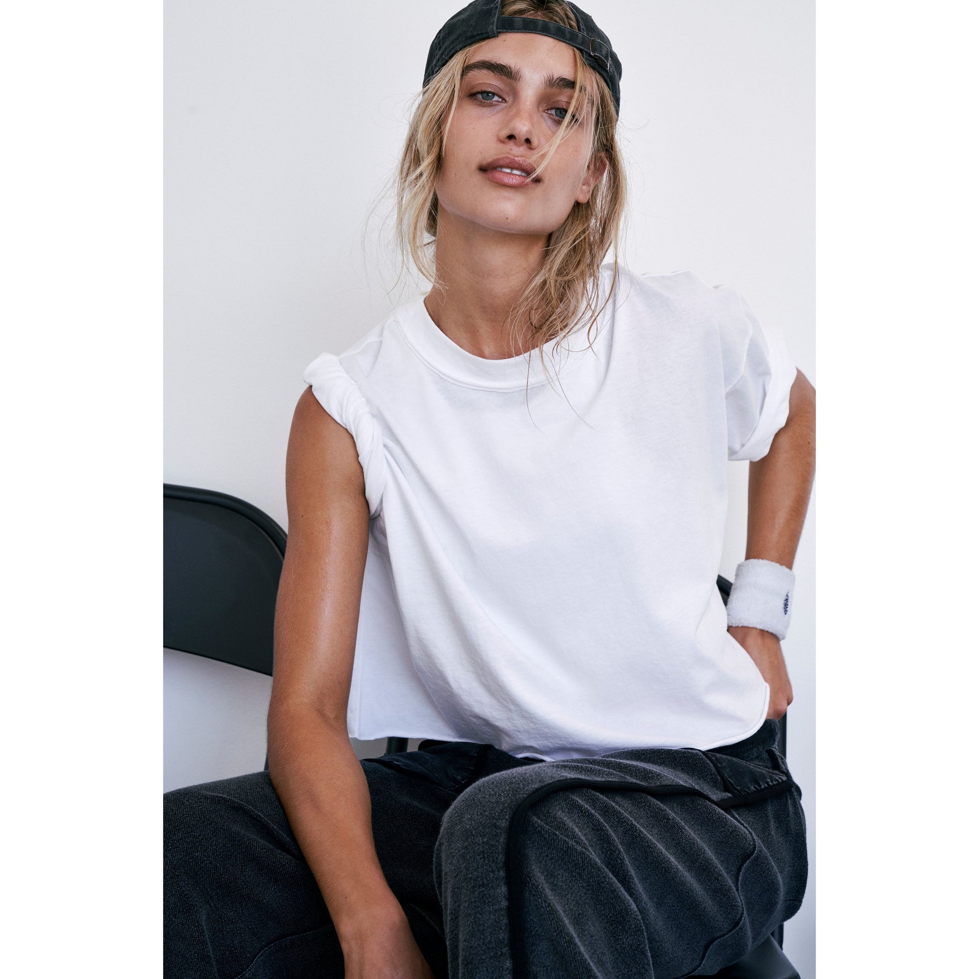 Young woman in an oversized white Free People Movement Inspire Tee and black cap sitting on a chair against a white background, looking casually at the camera.
