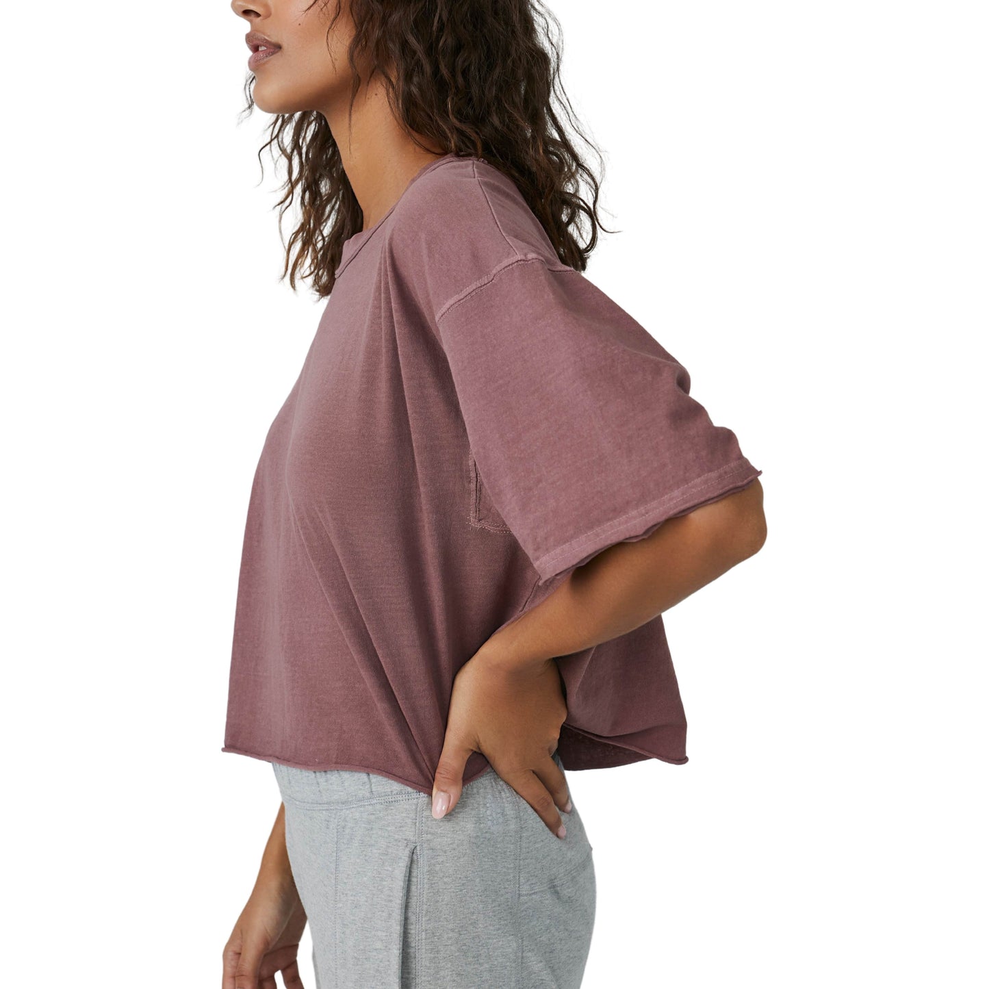 Side view of a woman wearing an oversized, dusty rose Free People Movement Inspire Tee and light gray sweatpants, with her hand partially tucked into her pocket.