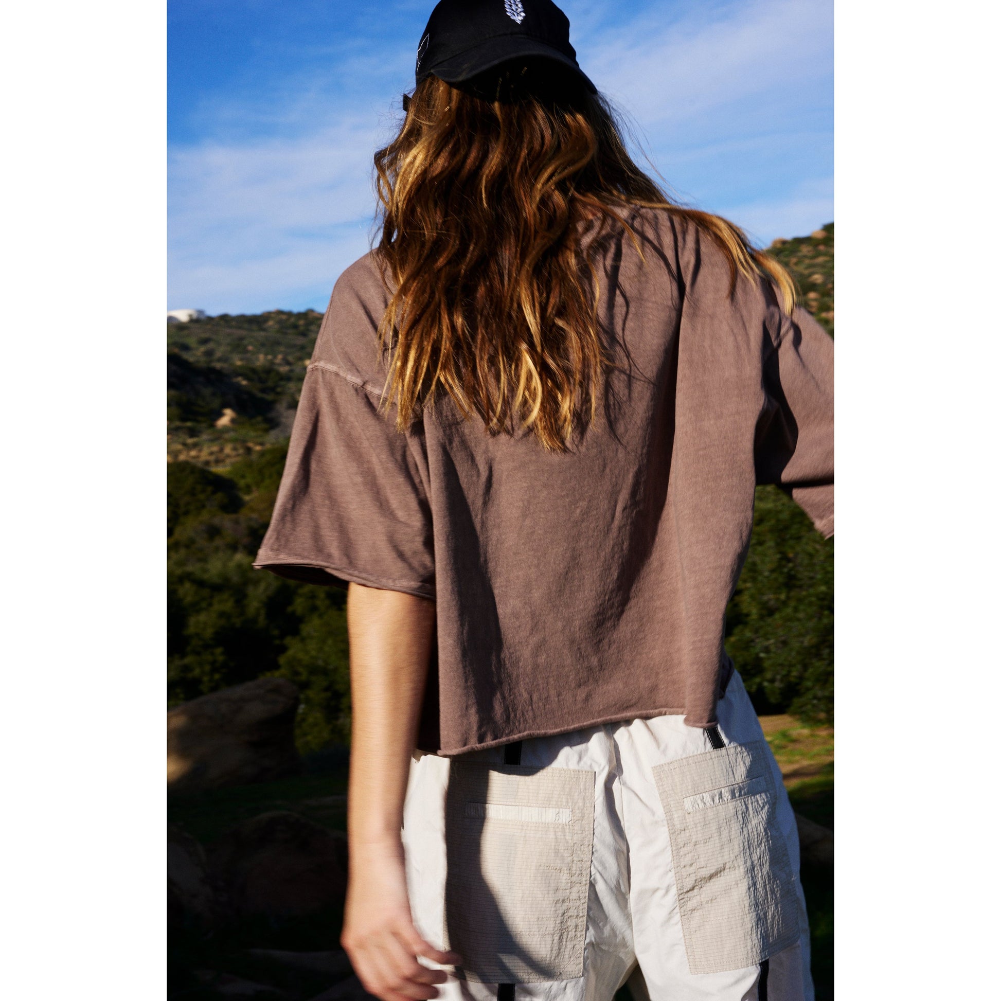 Woman with long hair standing outdoors, facing away, wearing a Wild Mustang Tee in a brown oversized boxy fit and white cargo pants, with a natural landscape in the background.