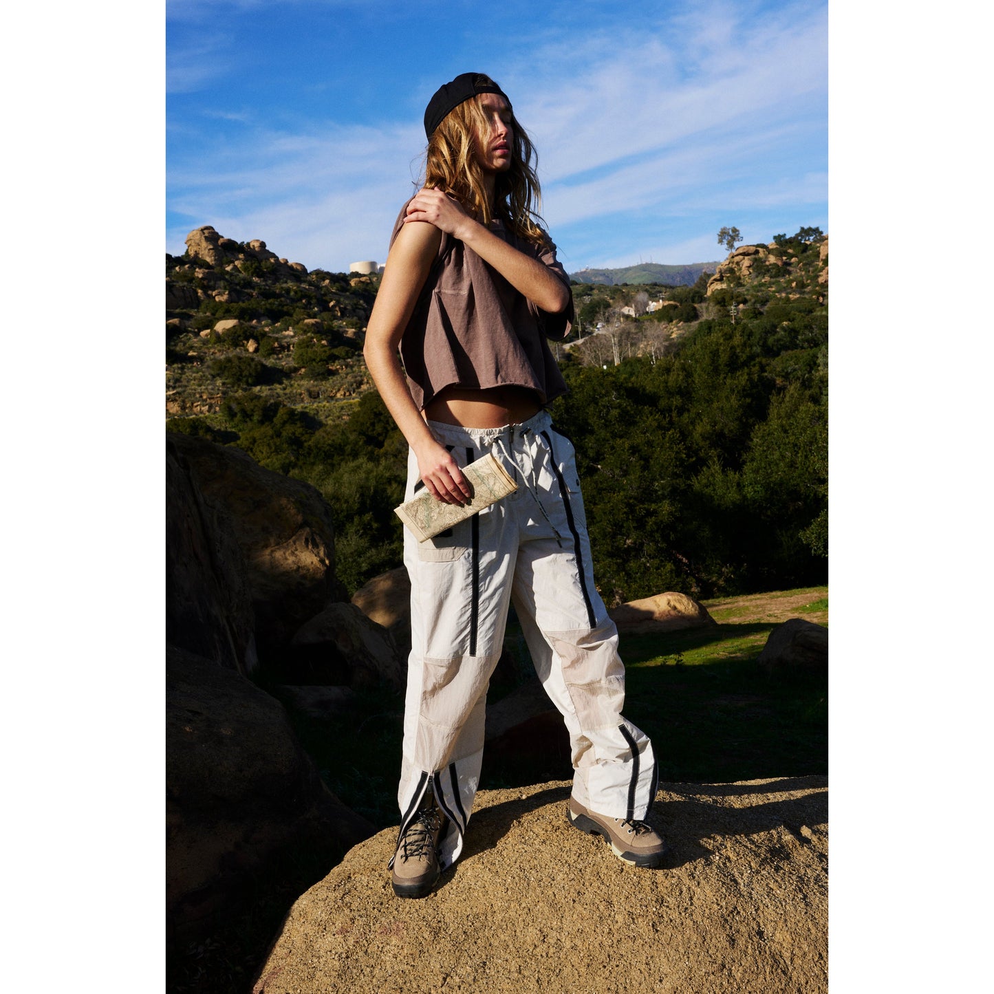 A person with long hair wearing a Free People Movement Inspire Tee in Wild Mustang, oversized boxy fit crop top, cargo pants, and boots stands holding a book in a rugged outdoor setting with hills in the background.