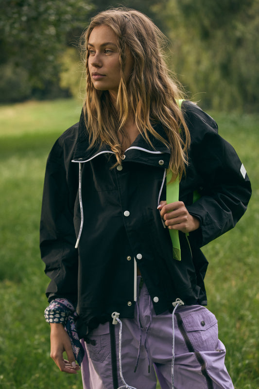 A young woman in a casual Rain & Shine Jacket in black by Free People Movement and lilac pants walks in a green park, looking thoughtfully to the side.