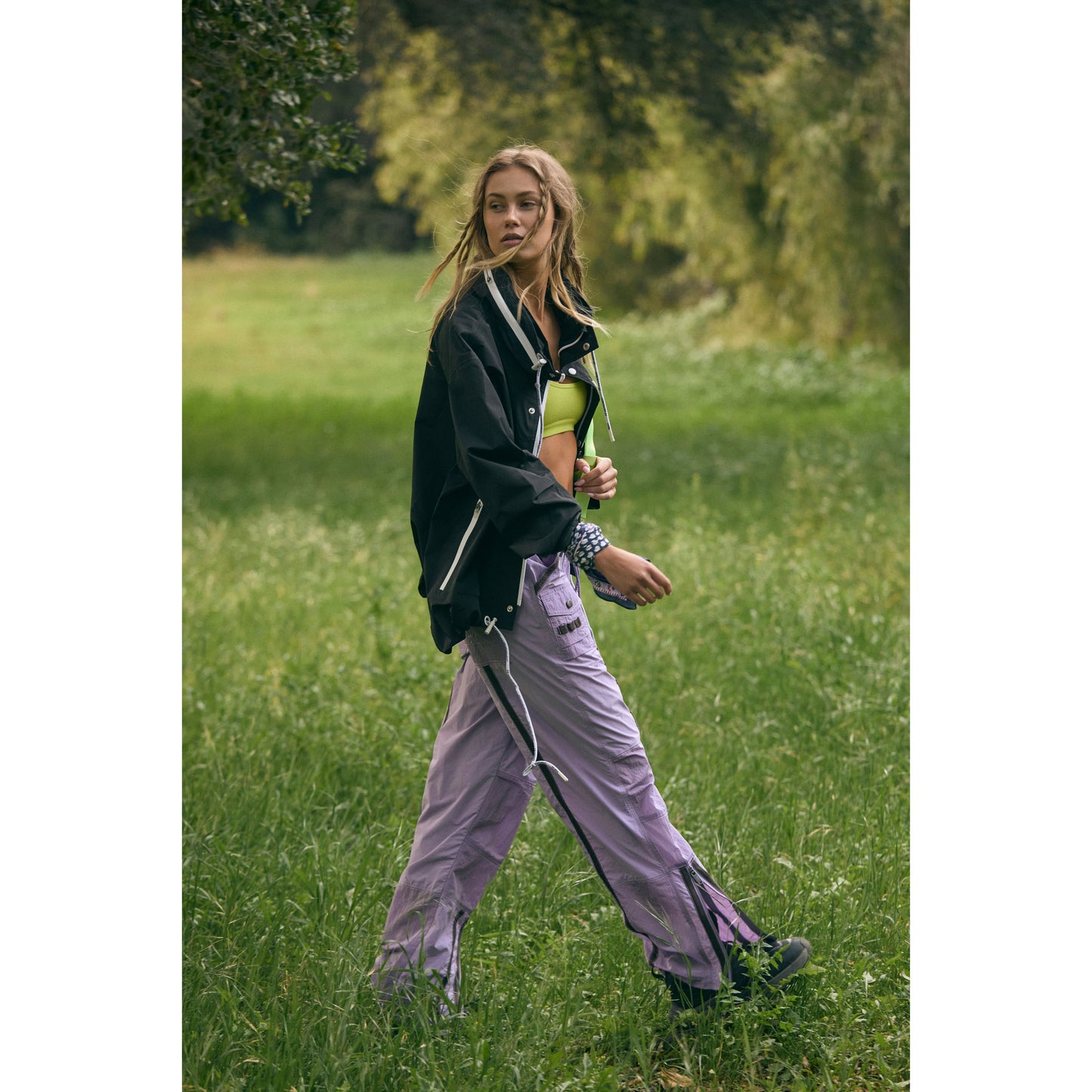Young woman walking in a grassy field, wearing a Free People Movement Rain & Shine Jacket in Black, purple pants, and holding a yellow book.