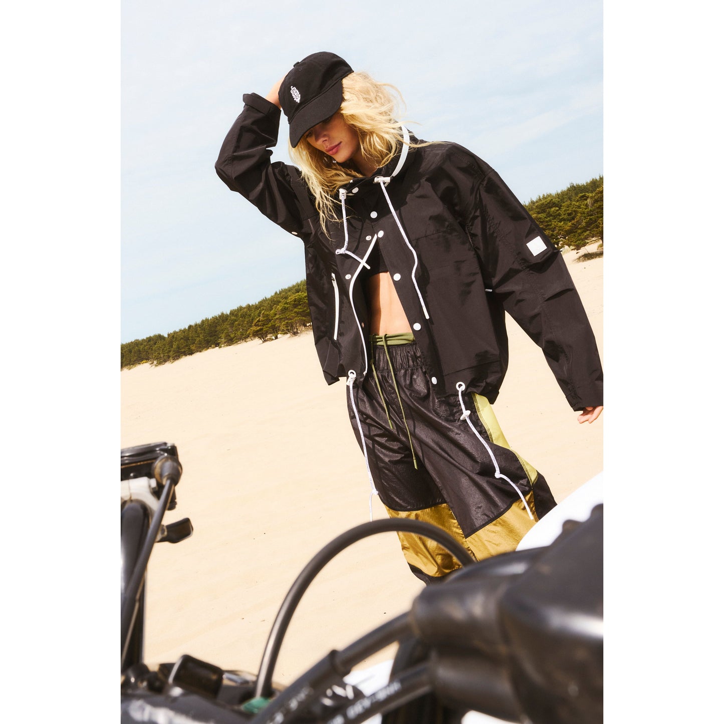 Woman in a stylish waterproof Free People Movement Rain & Shine Jacket in black and white and green pants posing near a motorcycle on a sandy beach.