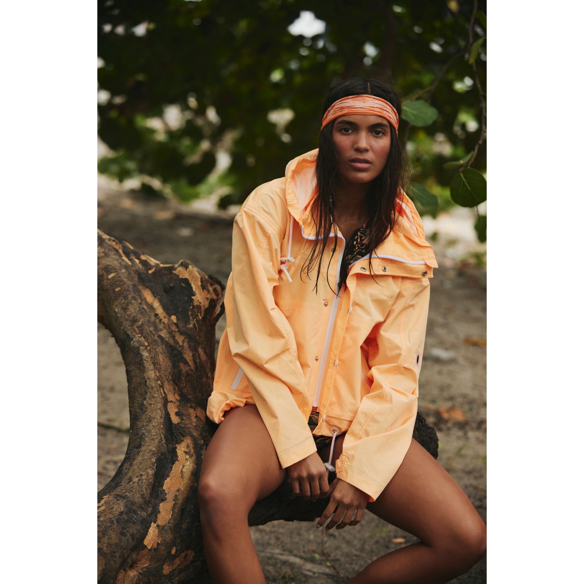 A woman in a Rain & Shine Jacket by Free People Movement and an orange headband sits on a gnarled tree trunk, looking away thoughtfully, surrounded by green foliage.
