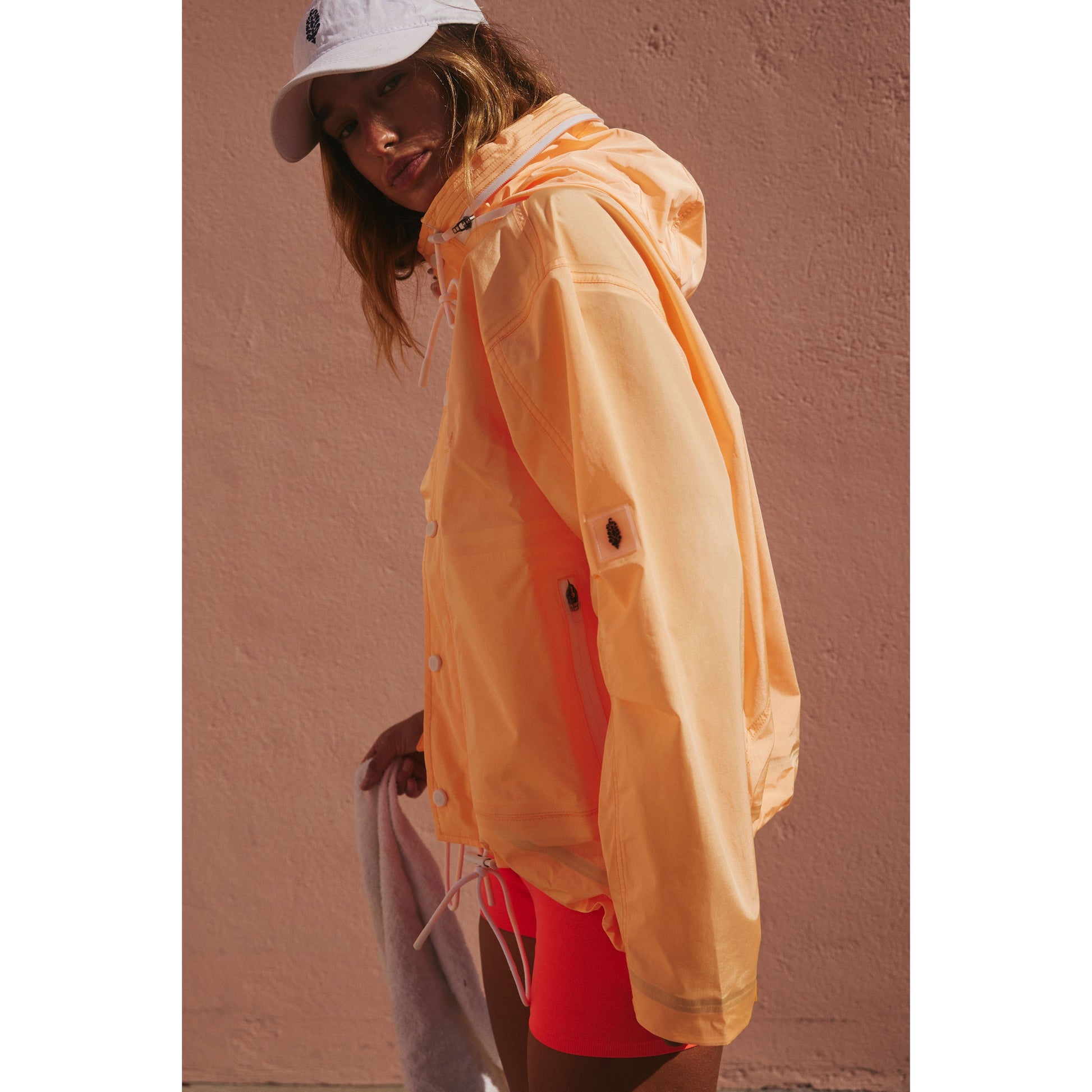 A woman models a vibrant orange Rain & Shine Jacket by Morning Sun and red shorts, paired with a white cap, against a pink wall. Brand Name: Free People Movement