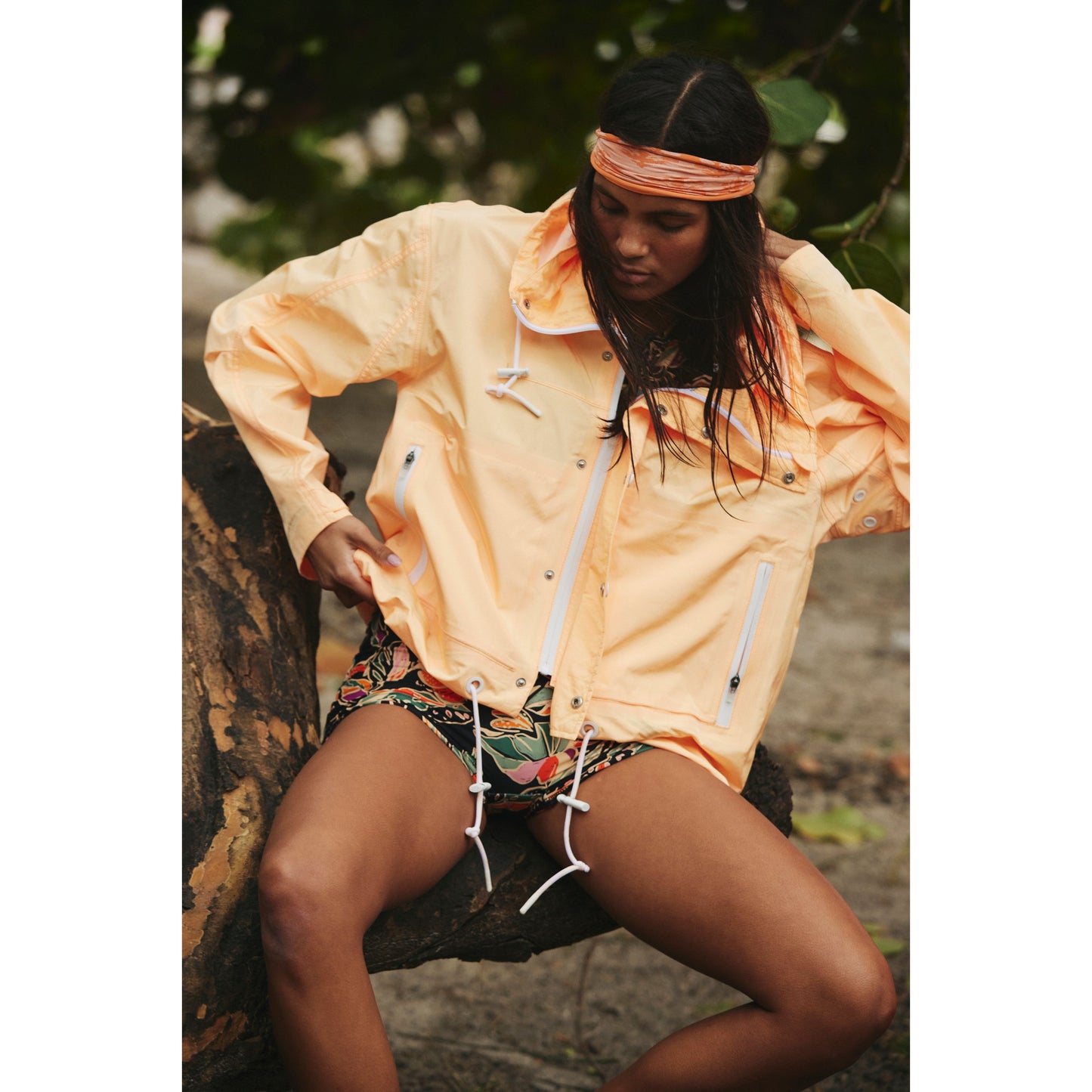 A woman sits on a log outdoors, wearing a Free People Movement Rain & Shine Jacket and patterned shorts, with a headband tied around her forehead.