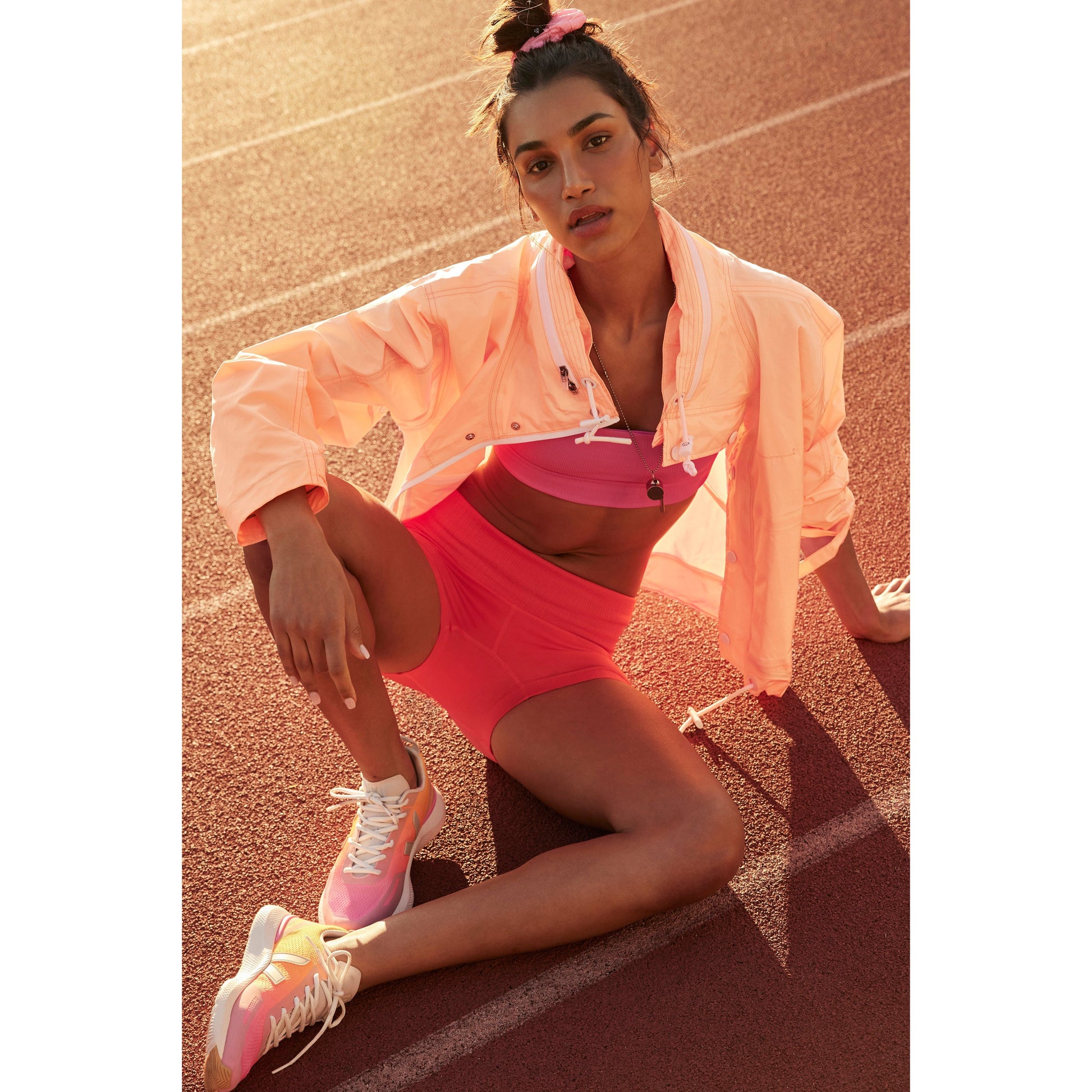 A woman in sportswear, sitting on a running track, wearing a Rain & Shine Jacket in Morning Sun color, shorts, and sneakers, looking up towards the sun.