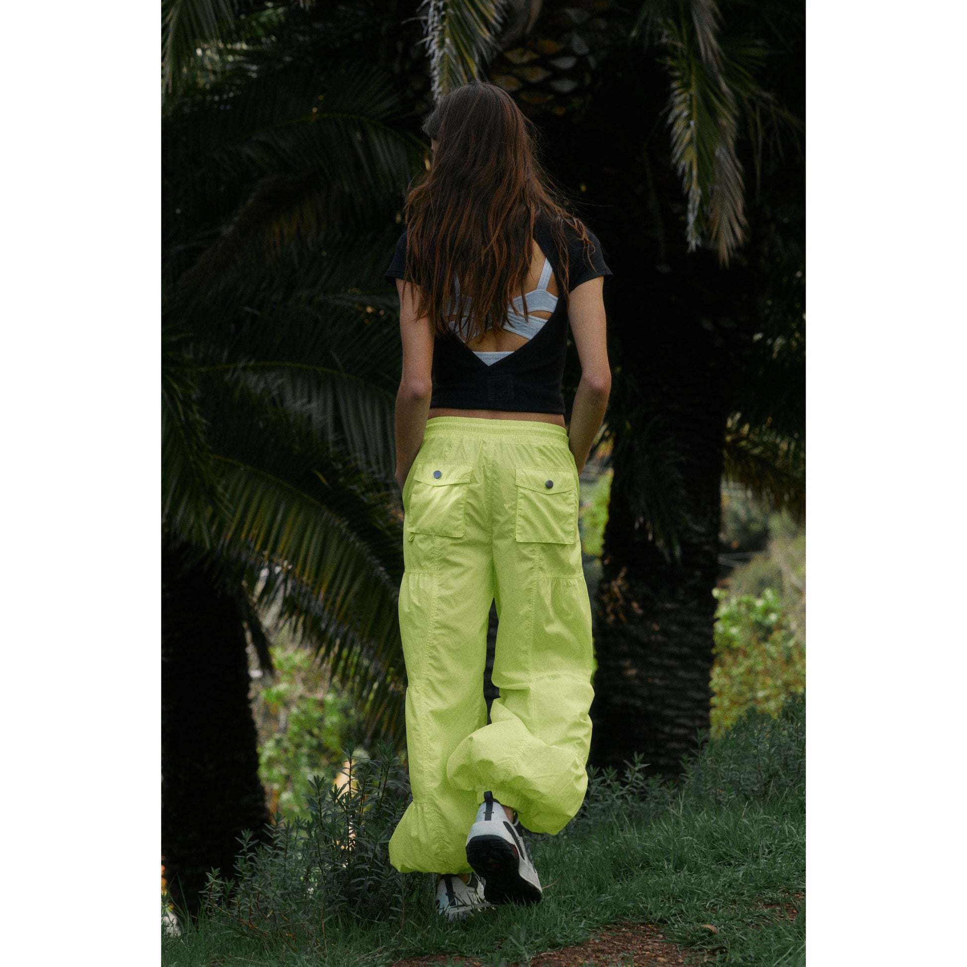 Woman from behind wearing Free People Movement's Lime Set Me Free Pant and a black and white top, standing amidst tropical foliage.