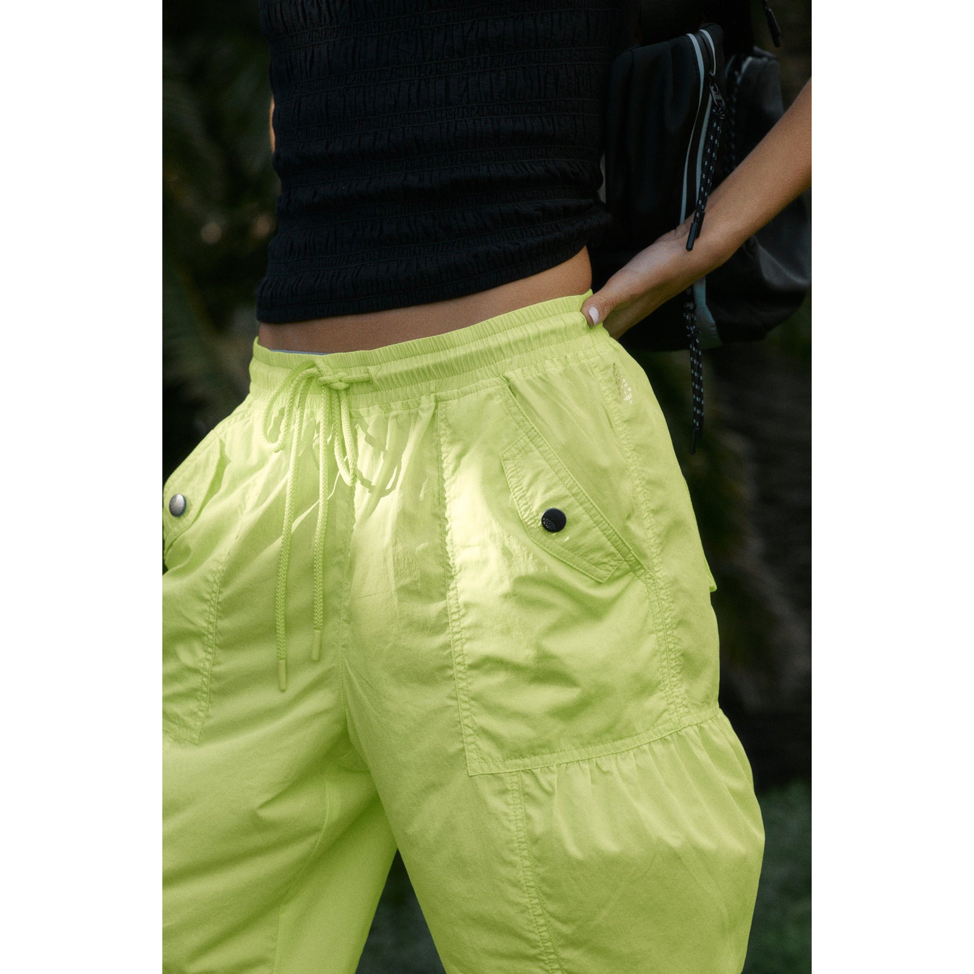 Close-up of a person wearing Free People Movement's Set Me Free Pant in Lime with drawstring waist, focusing on the midsection and hands in a tropical setting.