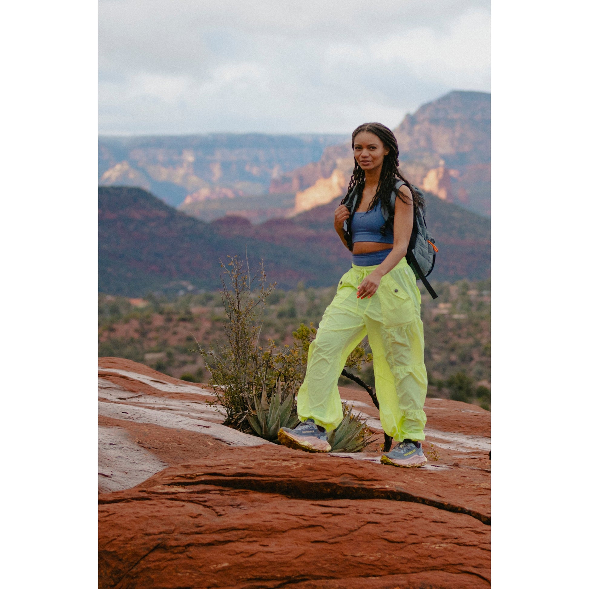 A woman in Free People Movement's Set Me Free Pant in Lime and a backpack standing on a red rocky trail, with mountains in the background.