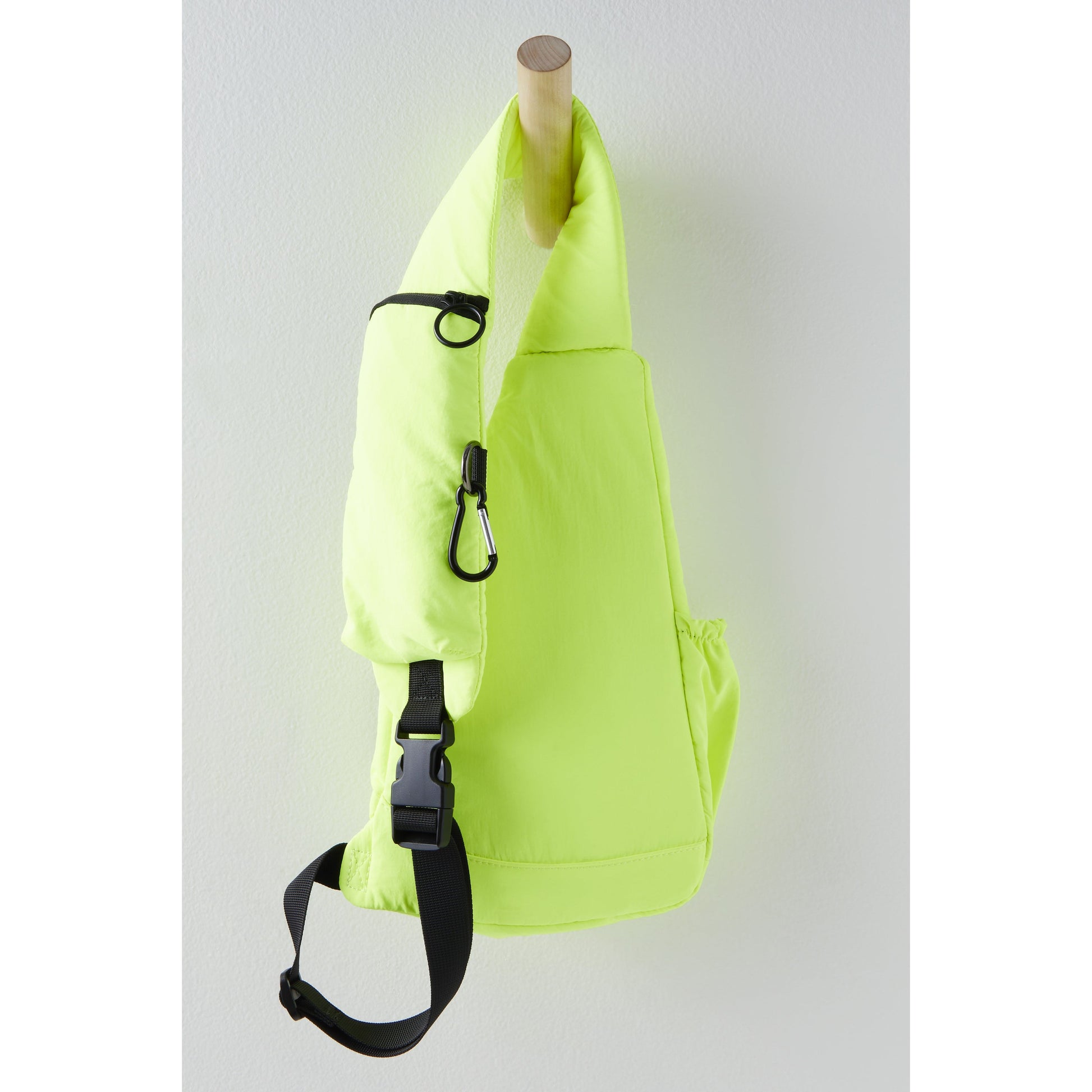 Neon green Renegade Sling bag with a black adjustable crossbody strap and carabiner, hanging on a wooden peg against a white background by Free People Movement.