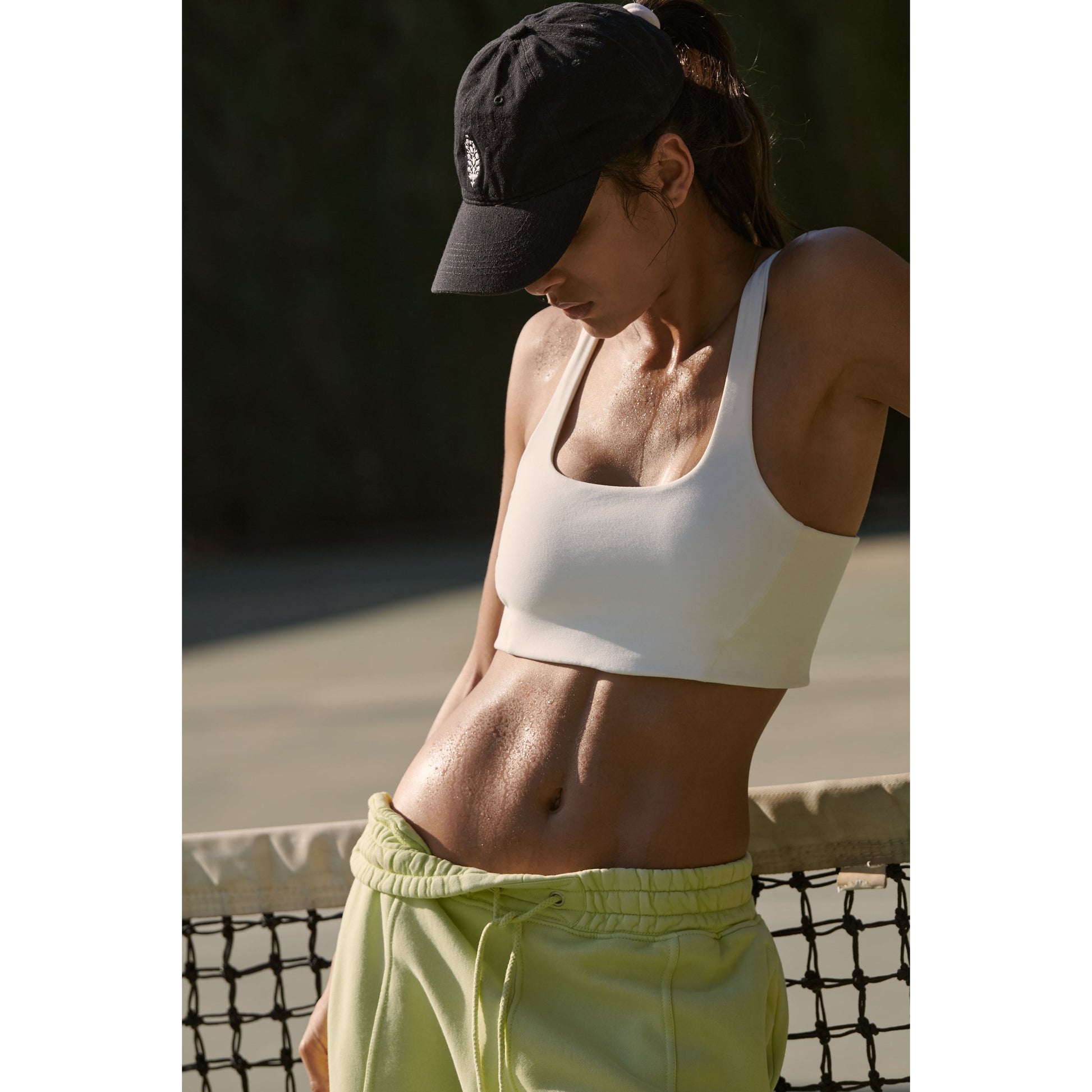 A woman in sportswear, including a Free People Movement Never Better SQ Neck Bra in white and yellow shorts, leaning on a tennis net, visibly perspiring, in a sunlit outdoor setting.