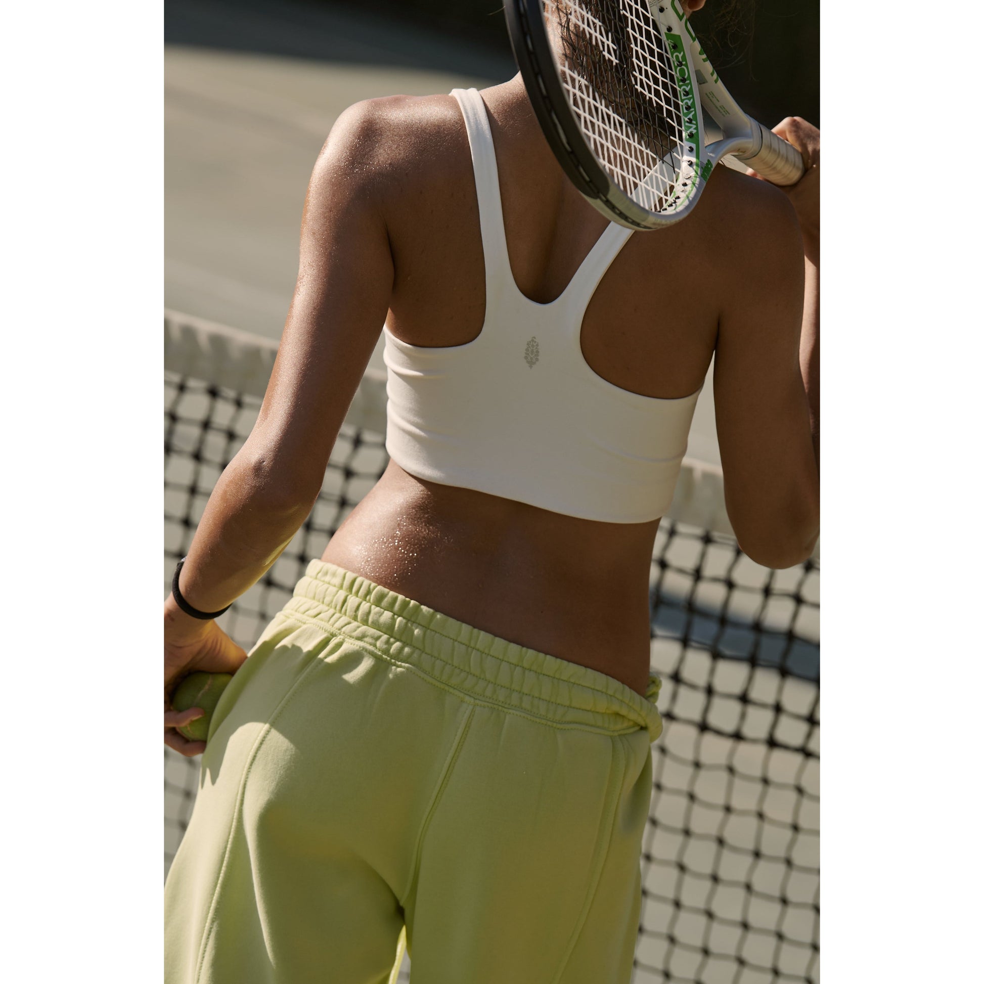 Woman in a Free People Movement Never Better SQ Neck Bra, White, and light green sweatpants holding a tennis racket, seen from the back.