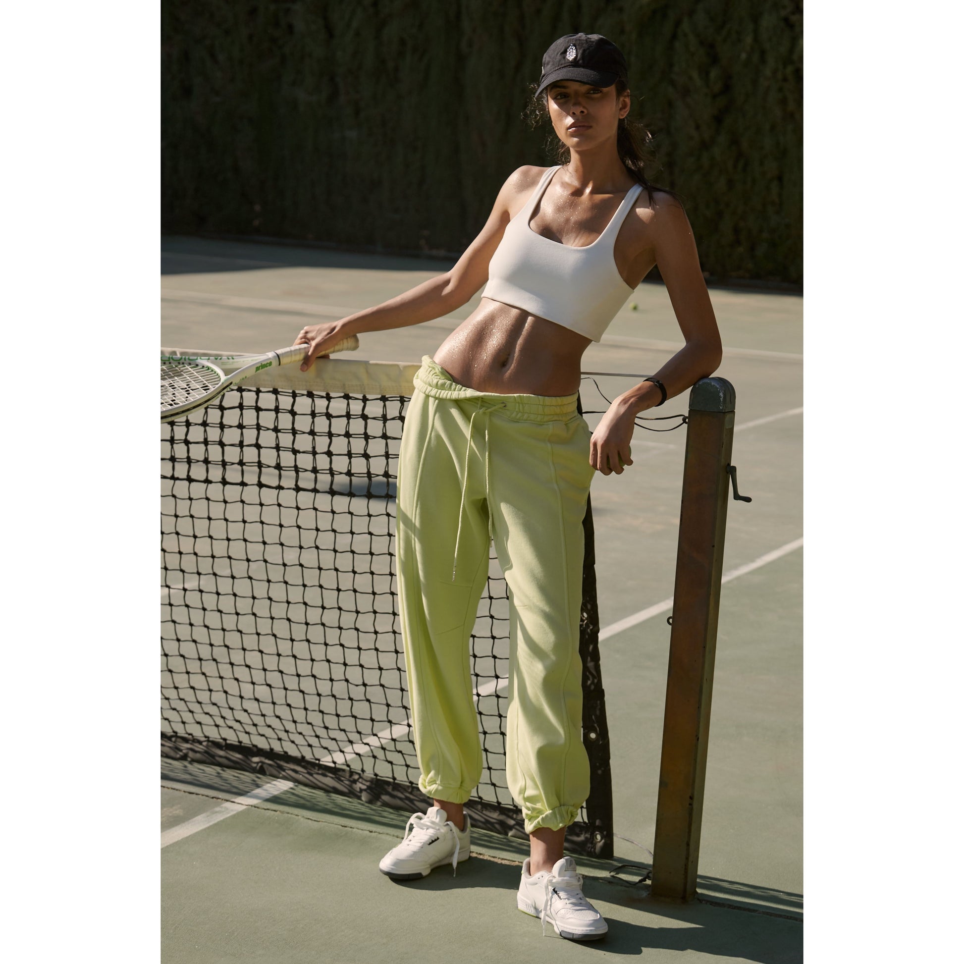 A woman in sporty attire stands beside a tennis net, holding a racket, wearing the Free People Movement Never Better SQ Neck Bra in White, lime green track pants, and a black cap.