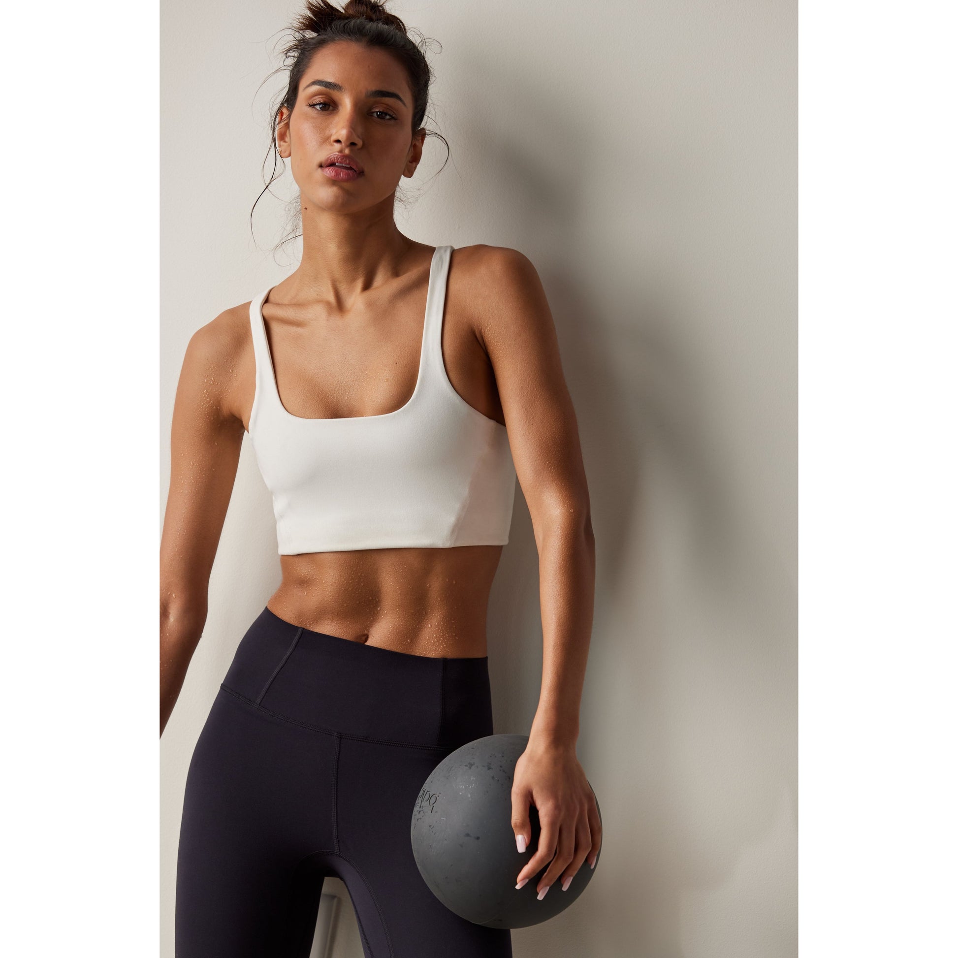 A woman in athletic wear, featuring a Free People Movement Never Better SQ Neck Bra in White with a breathable design, holds a medicine ball against her hip in a neutral-toned studio.