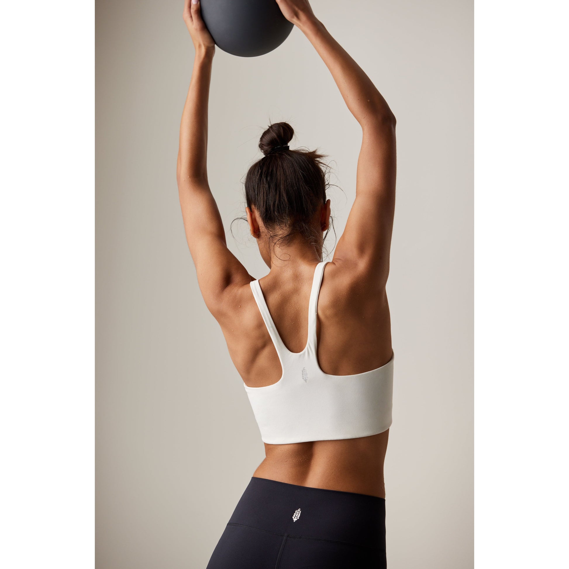 A woman in a Free People Movement Never Better SQ Neck Bra in White and black leggings, viewed from behind, lifting a medicine ball above her head against a neutral background.
