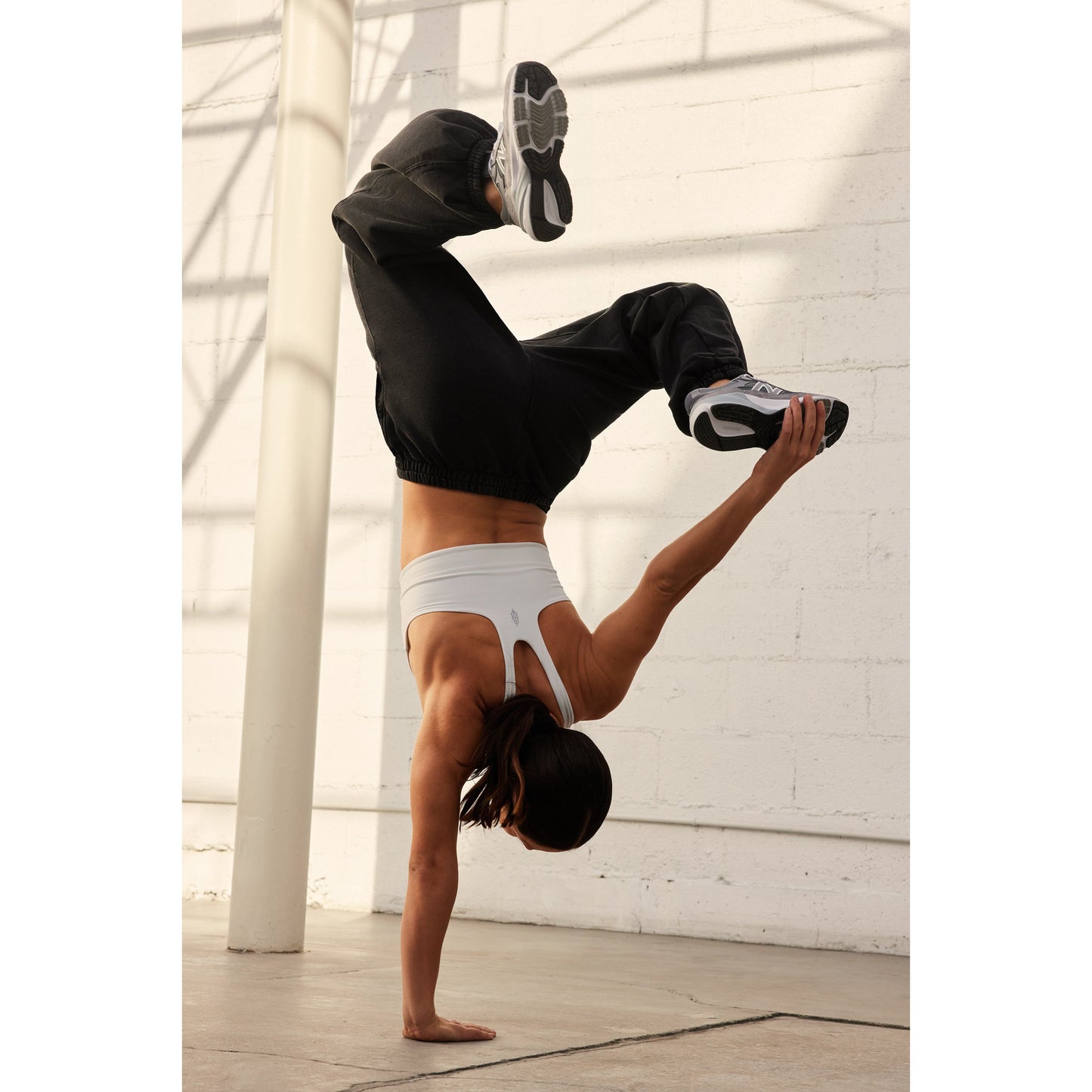A woman performs a handstand in an urban setting, wearing a white Never Better SQ Neck Bra by Free People Movement, black pants, and sneakers.