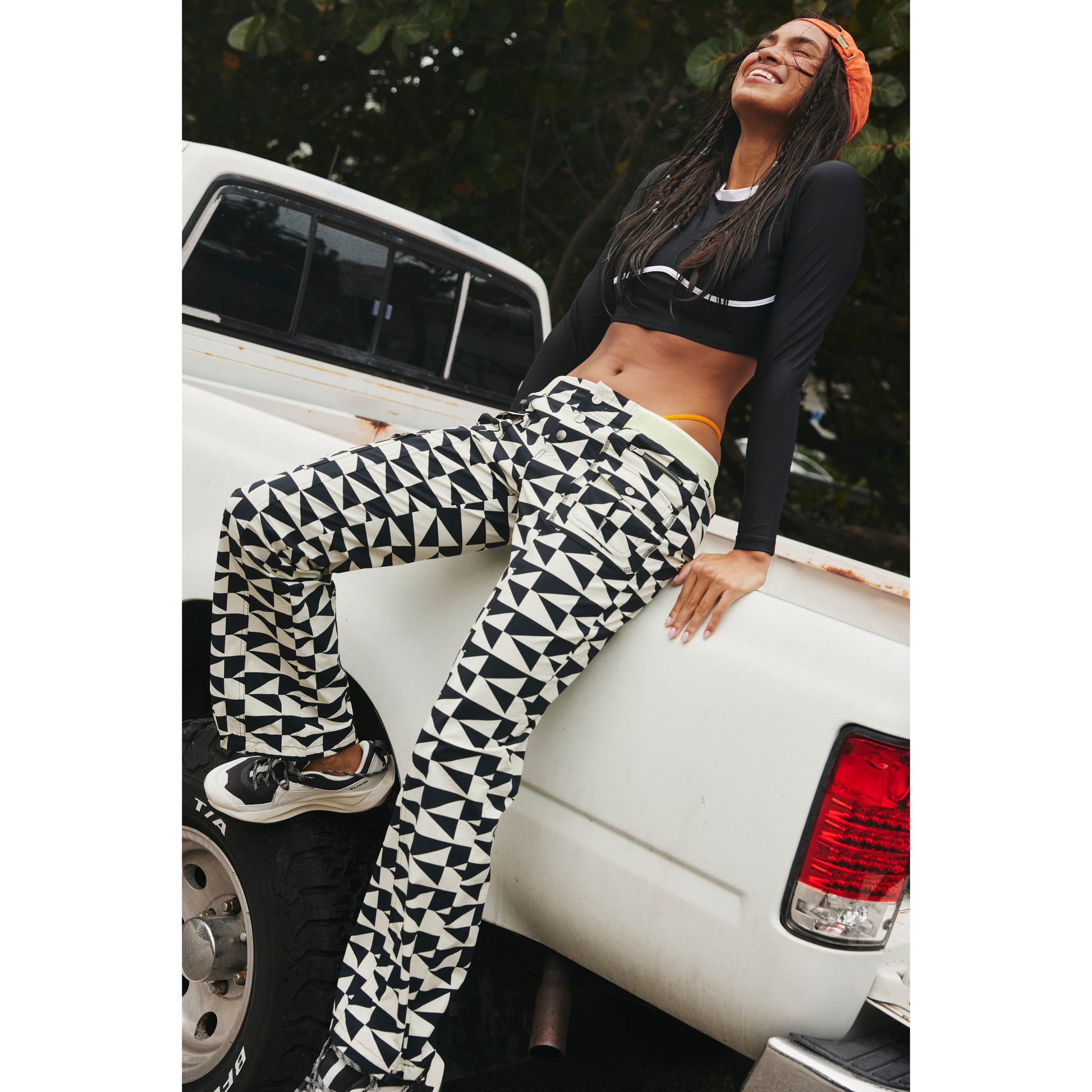 A joyful woman wearing a Free People Movement Printed Cascade Flare headband, bralette, and Off The Grid Combo pants sits on the tailgate of a white pickup truck.