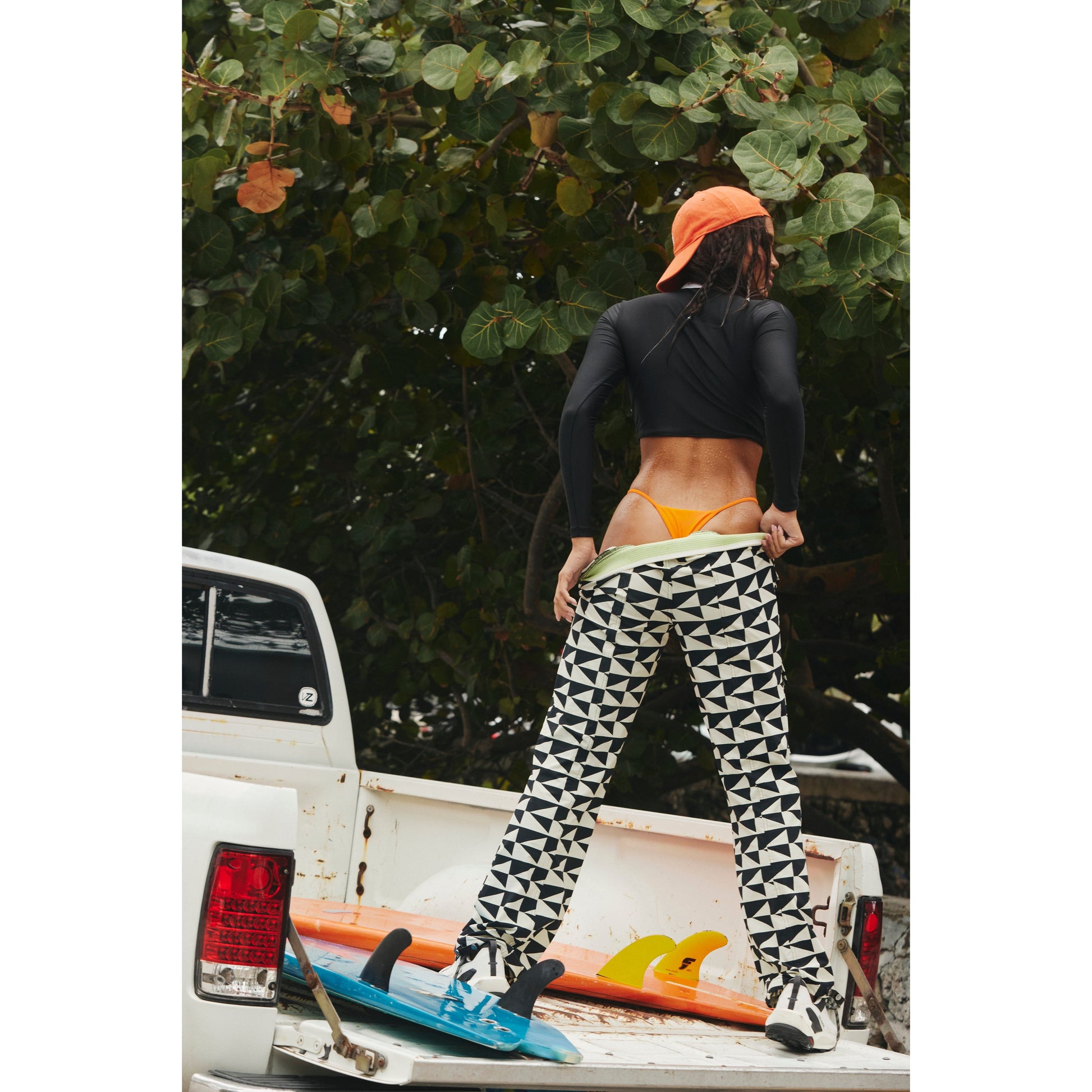 Woman in a black top and Free People Movement's Printed Cascade Flare and Off The Grid Combo pants standing on the back of a pickup truck with a surfboard, facing a tree.