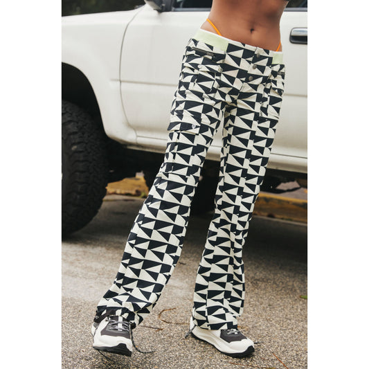 A person stands beside a white pickup truck, wearing Free People Movement's Printed Cascade Flare pants in the Off The Grid Combo pattern and sneakers. Only the torso and legs are visible.