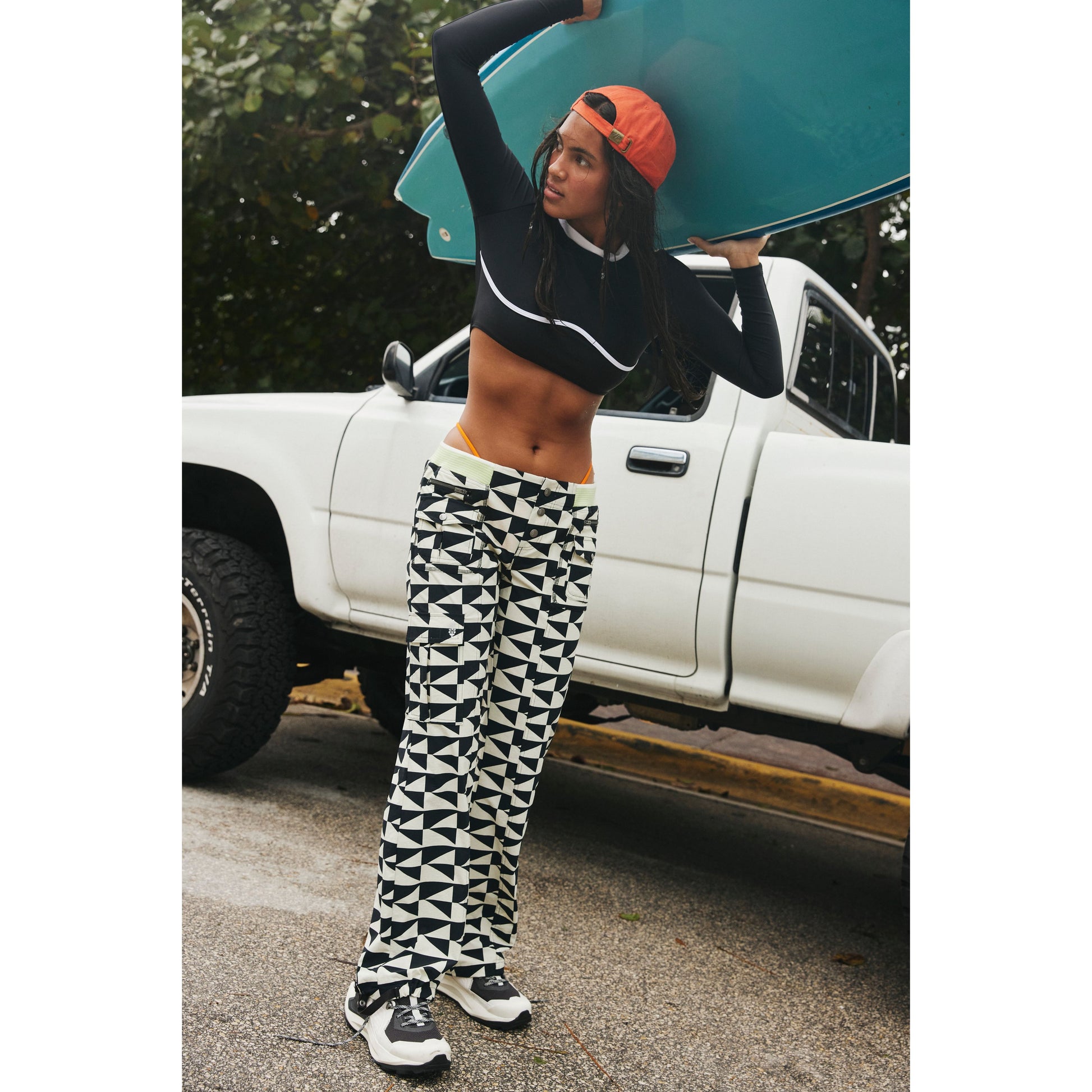 A woman in a Free People Movement sports bra and Off The Grid Combo patterned pants carries a blue surfboard beside a white pickup truck.