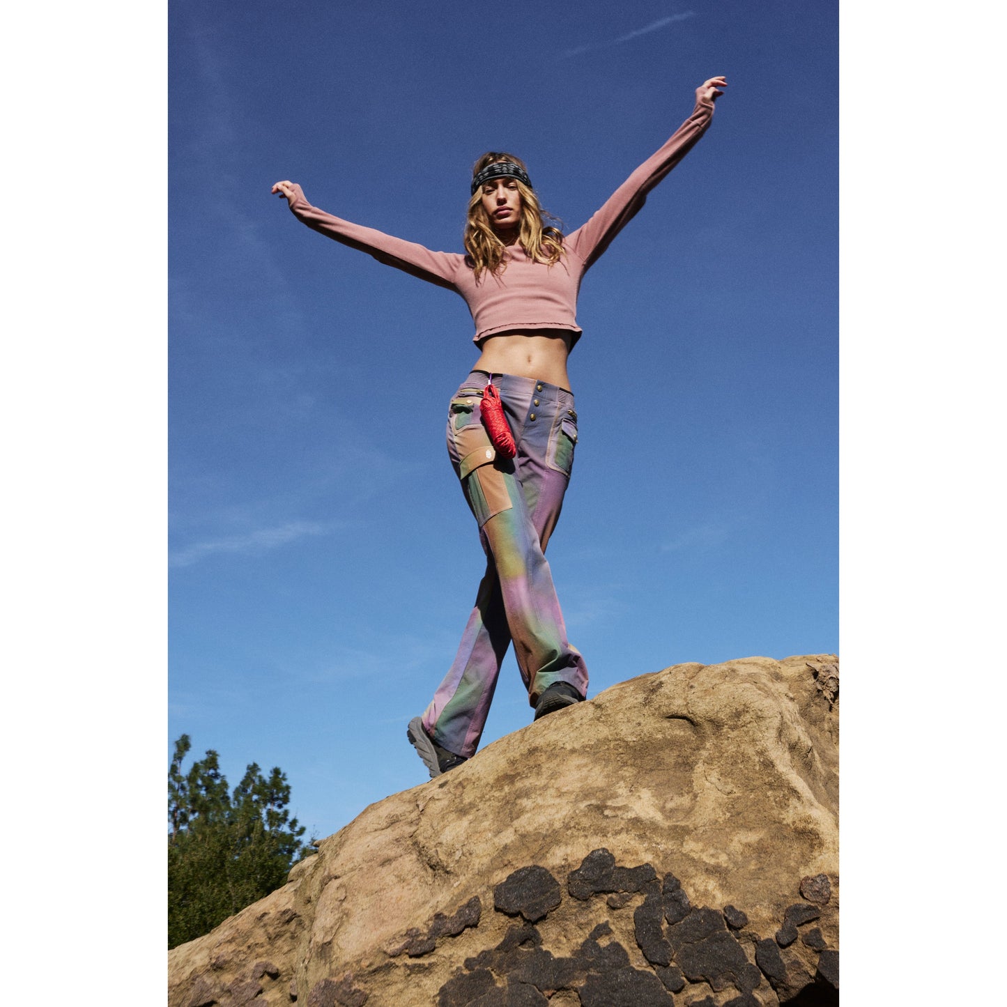 A woman in a pink crop top and Free People Movement Galaxy Gradient Printed Cascade Flare pants stands triumphantly on a rocky hill against a clear blue sky.