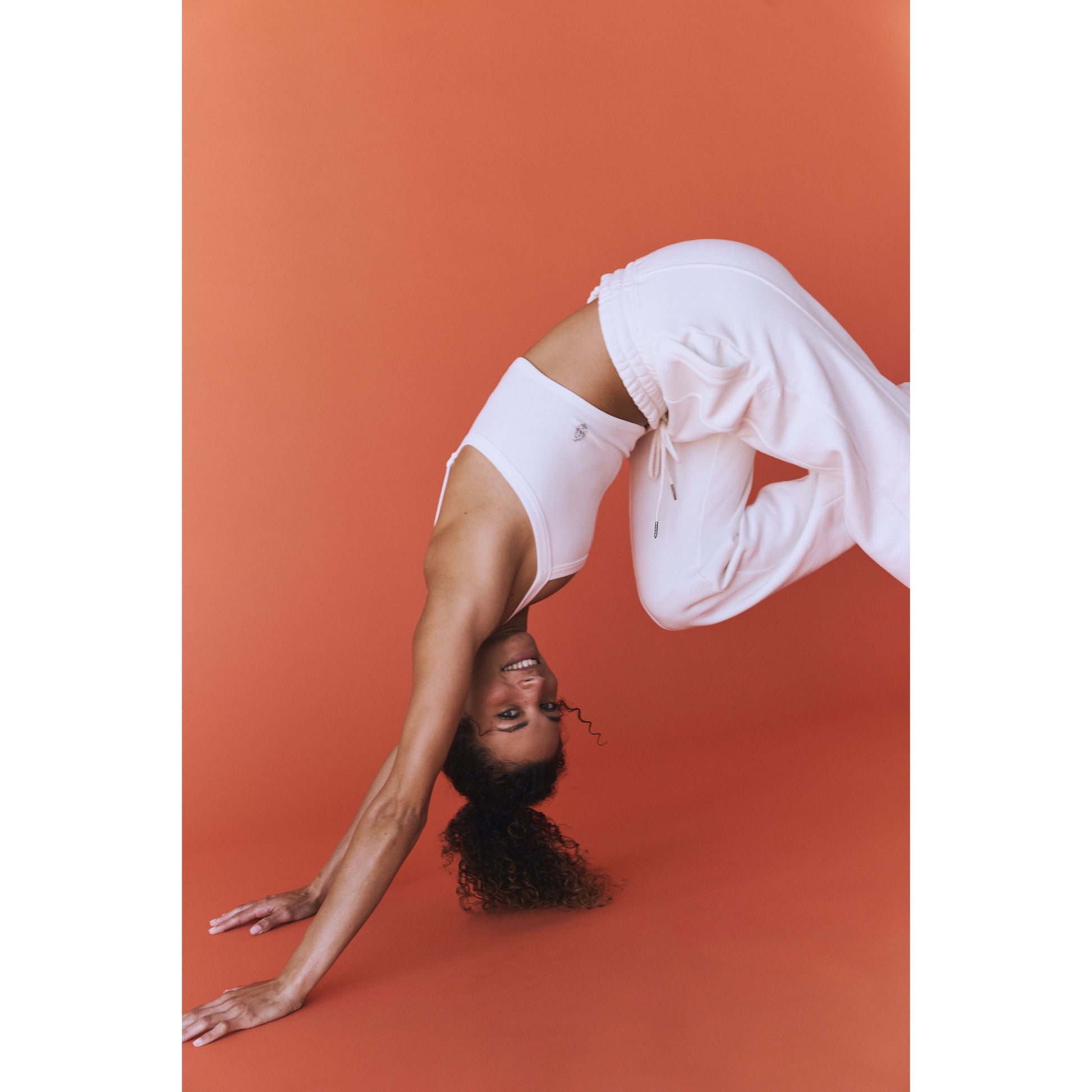 A woman performs a backbend in a Free People Movement All Clear Cami Solid, White outfit against an orange background, smiling at the camera.