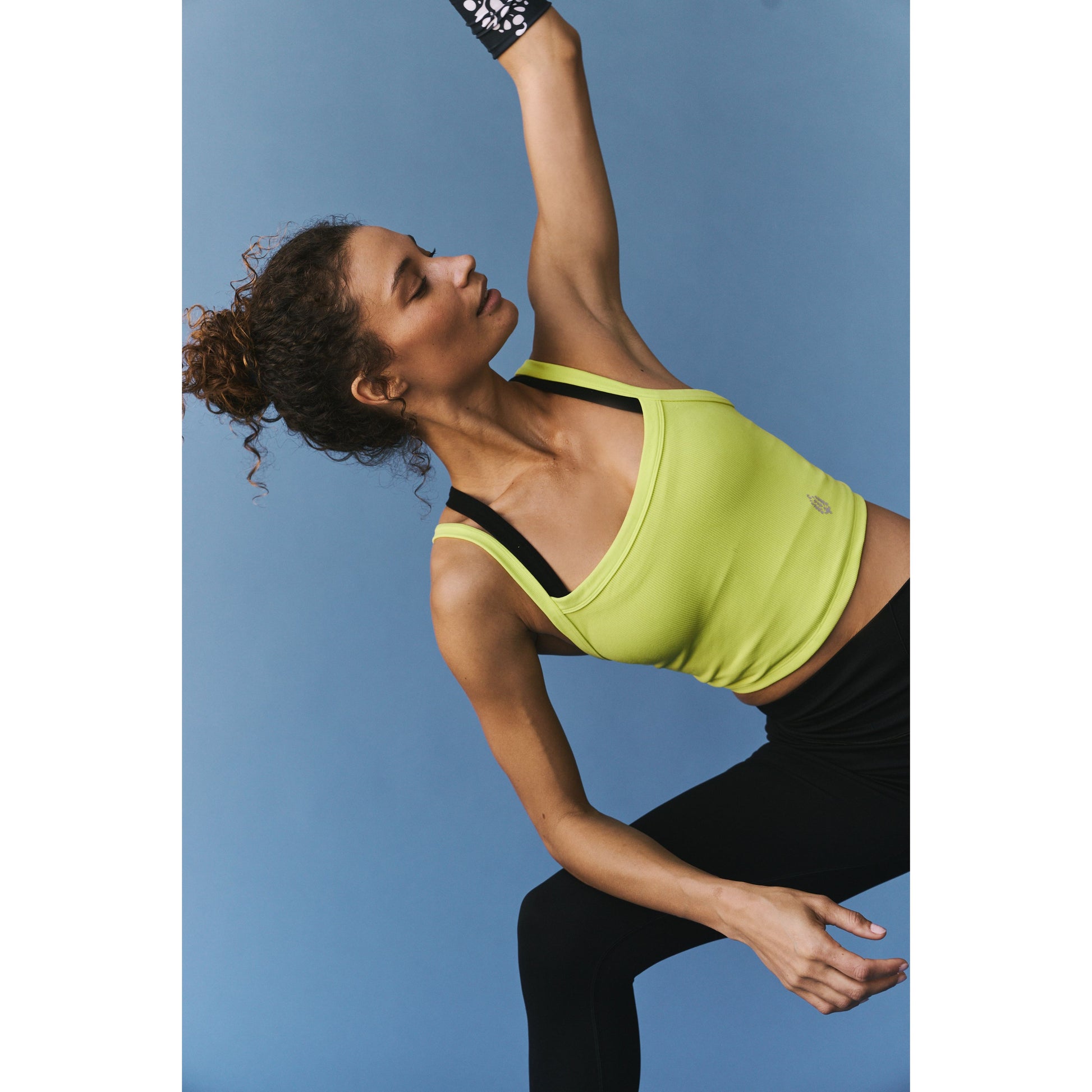 A woman in a Free People Movement lime green All Clear Cami Solid tank top and black leggings stretches her arm upwards while looking focused against a blue background.