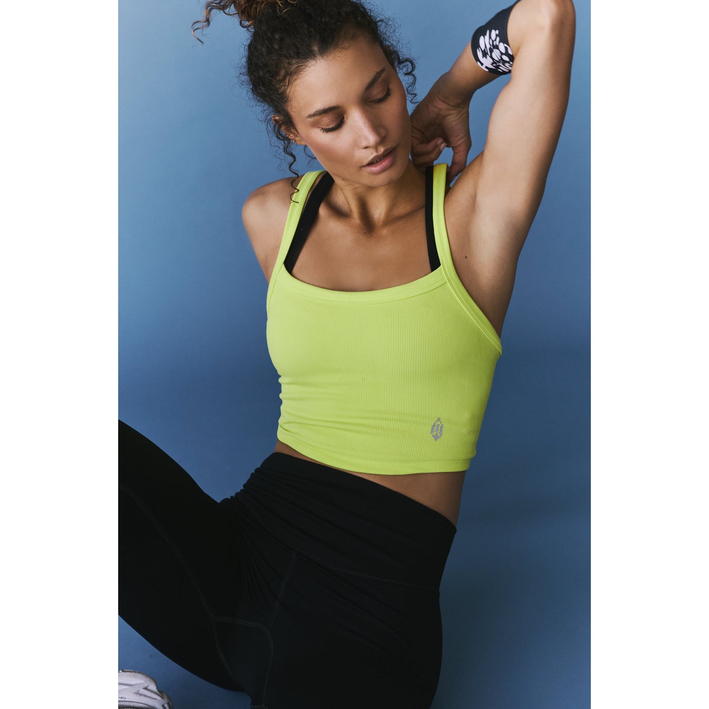 A woman in a Free People Movement All Clear Cami Solid in Highlighter Yellow and black leggings stretching her arm behind her head against a blue background.