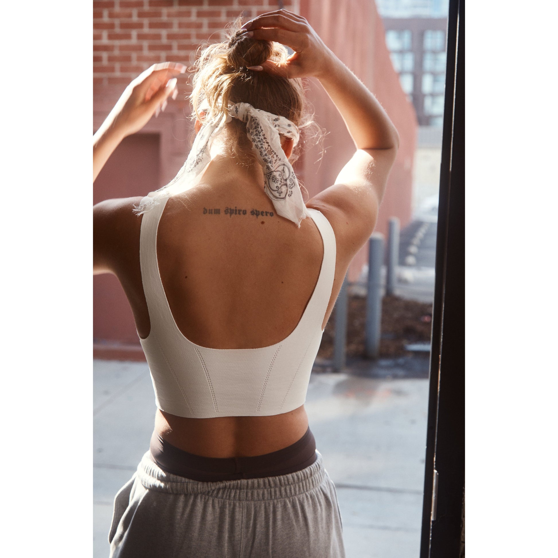 Woman tying her hair, viewed from behind, with a tattoo reading "dum spiro spero" on her upper back. She wears a Free People Movement Stong Core Corset Cami in White and gray pants.