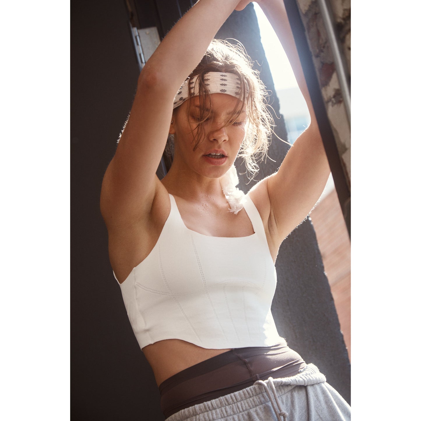 A woman in a Free People Movement Stong Core Corset Cami in White, with workout gear and a headband, tying her hair up under sunlight, showing slight perspiration.