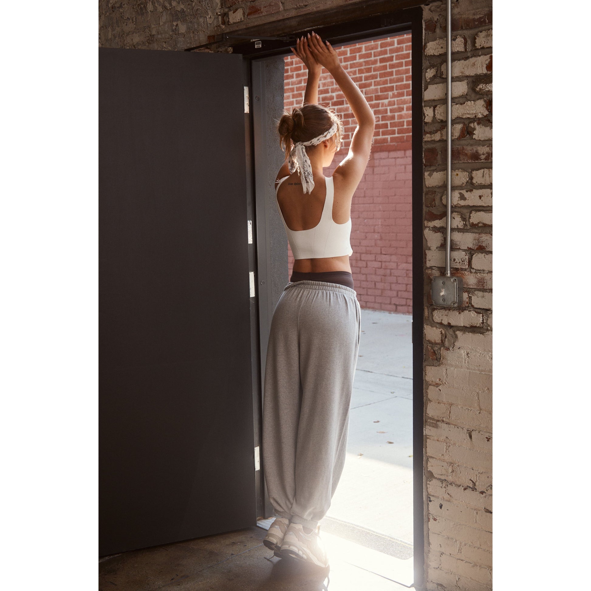 A woman in a Free People Movement Strong Core Corset Cami, White and grey sweatpants stretches her arms up near an open doorway, facing a brick wall.