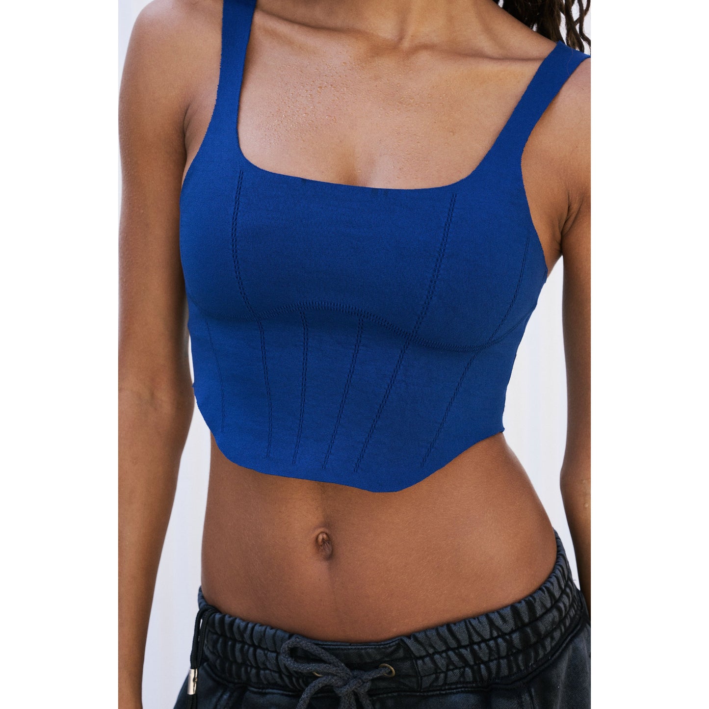 A close-up of a woman in a blue crop top with a scoop neckline and black drawstring pants, focusing on her torso wearing the Strong Core Corset Cami in Dark Purple by Free People Movement.