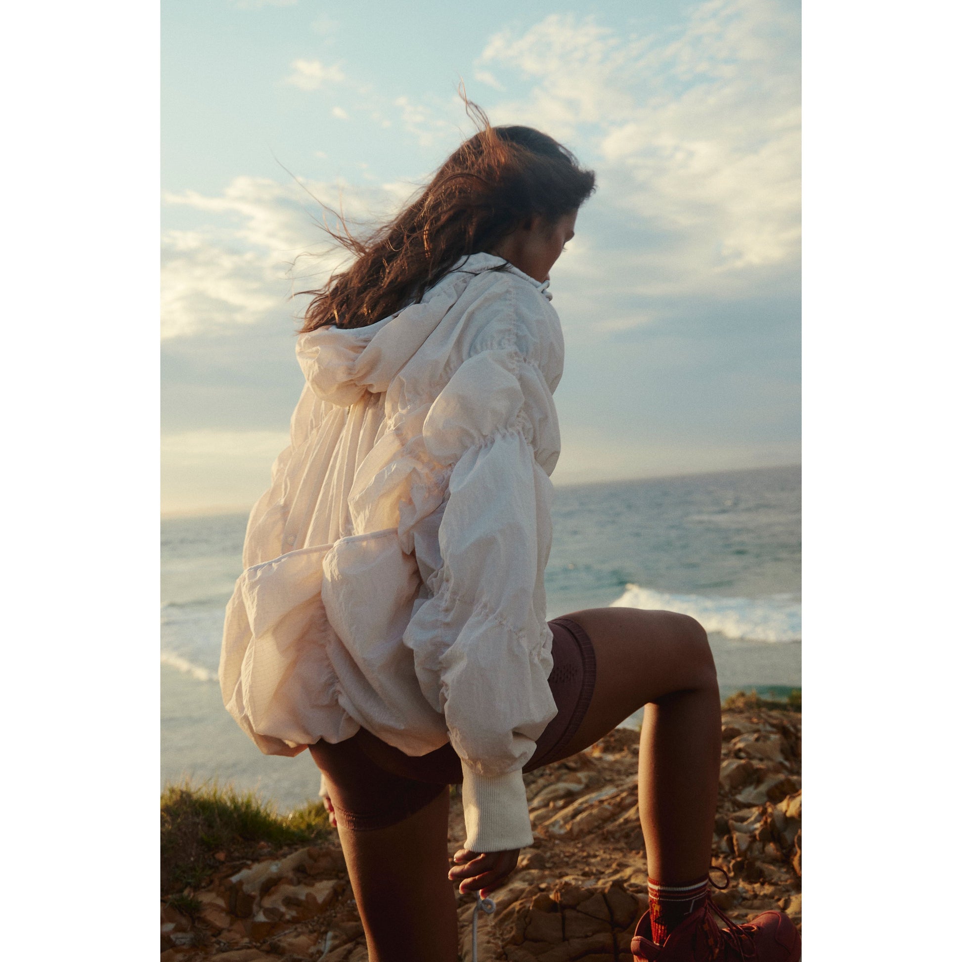 A woman in a white Free People Movement blouse and breathable nylon shorts stands on rocks near the sea, facing away, with wind blowing her hair wearing the Happy Camper Pullover in Bleached Clay.