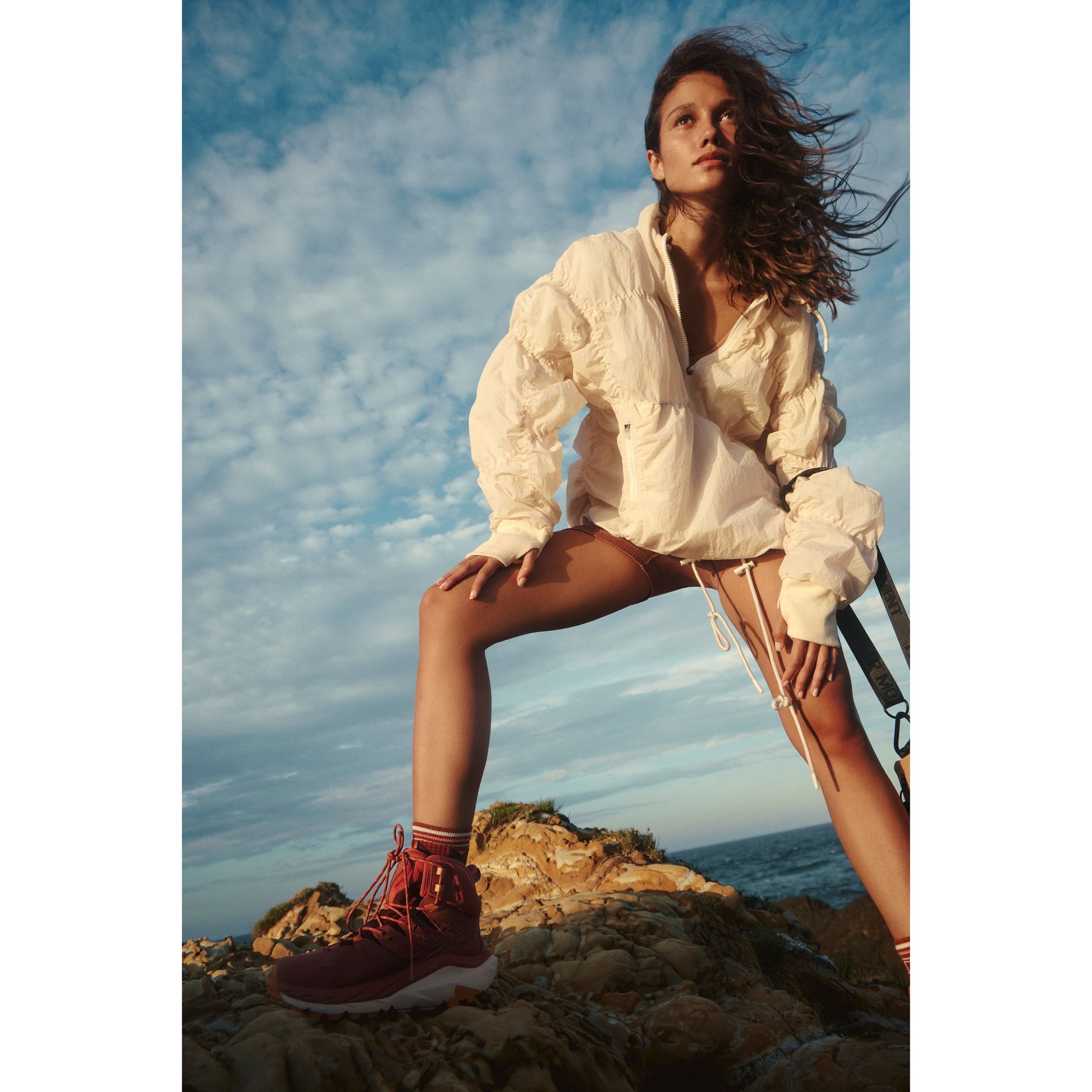 A young woman in a white blouse and red hiking boots, wearing a Free People Movement Happy Camper Pullover in Bleached Clay, poses on rocky coastal terrain under a clear sky.