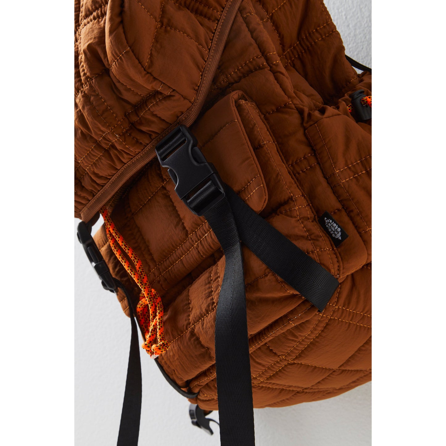 Close-up of a Free People Movement Summit Backpack in Brown with adjustable shoulder straps and a flap-top closure, featuring an orange and black rope detail.