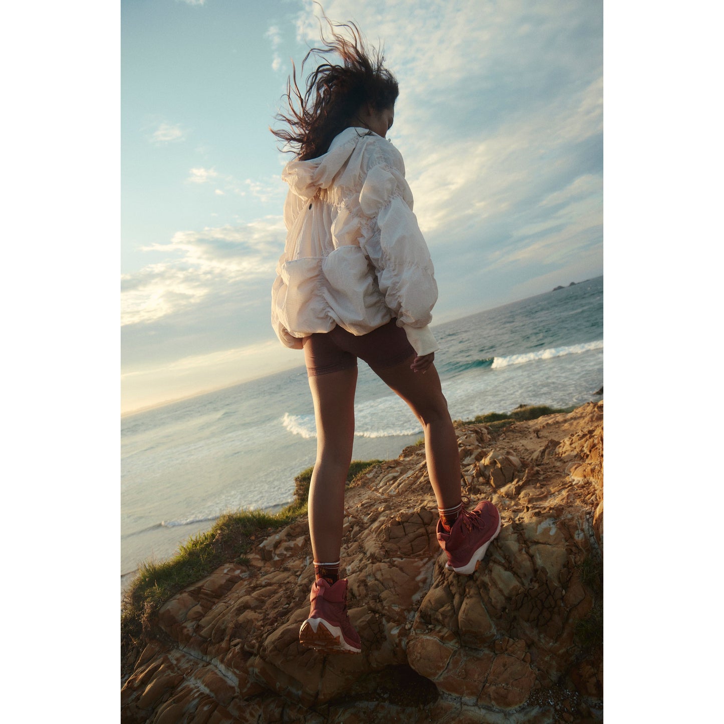 Woman in a Free People Movement Happy Camper Pullover in Bleached Clay and shorts jumping on a rocky shoreline, with the ocean in the background at sunset.