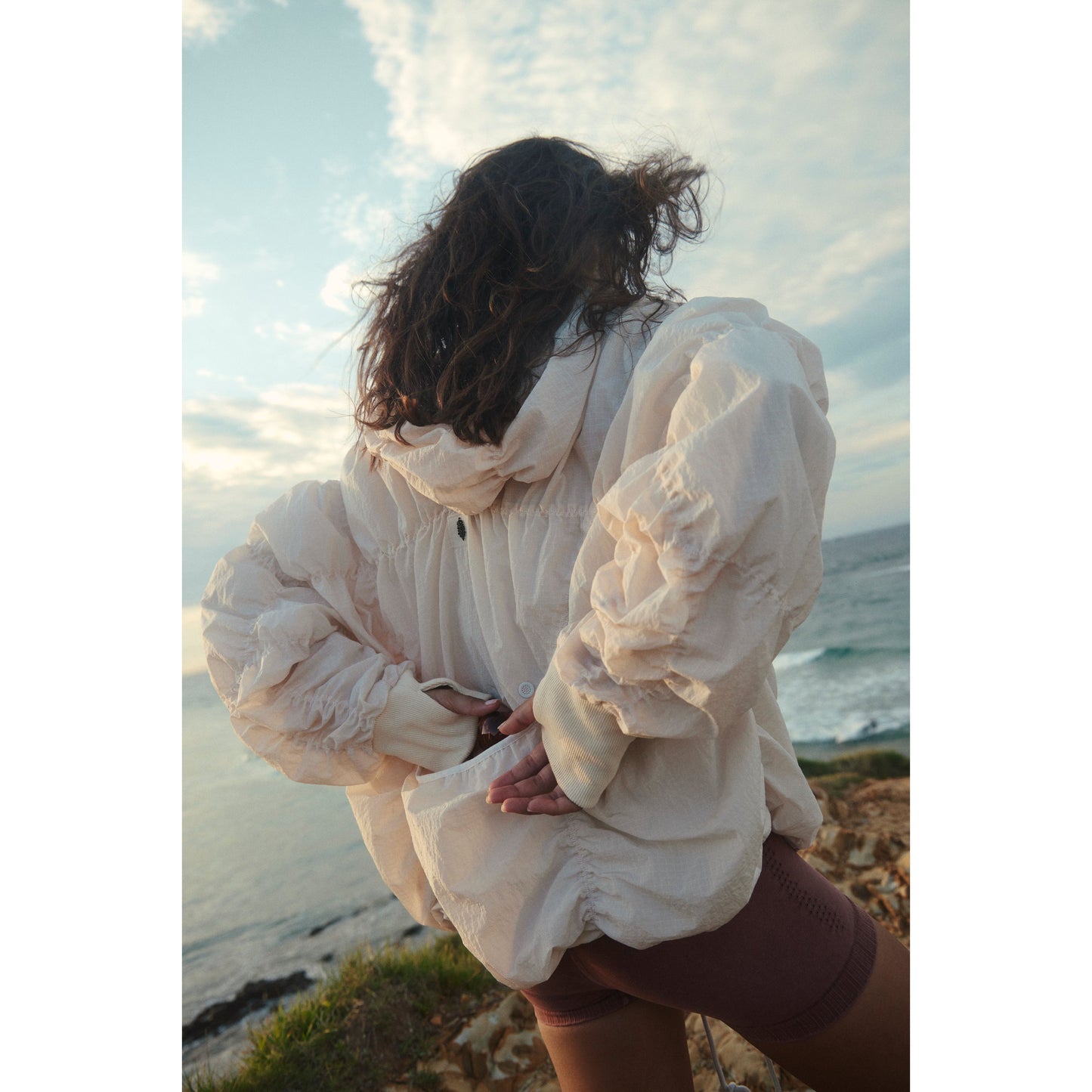 Woman in a Free People Movement Happy Camper Pullover in Bleached Clay stands facing the ocean, her curly hair blown by the wind, during sunset.