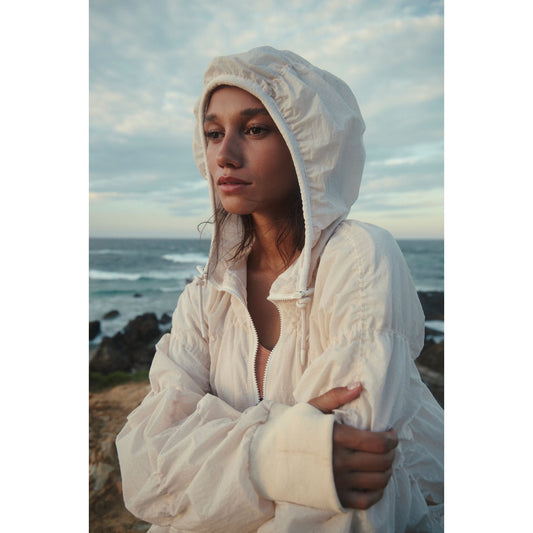 A woman in a white, wind- and water-resistant Happy Camper Pullover by Free People Movement stands by the sea at dusk, looking contemplative.