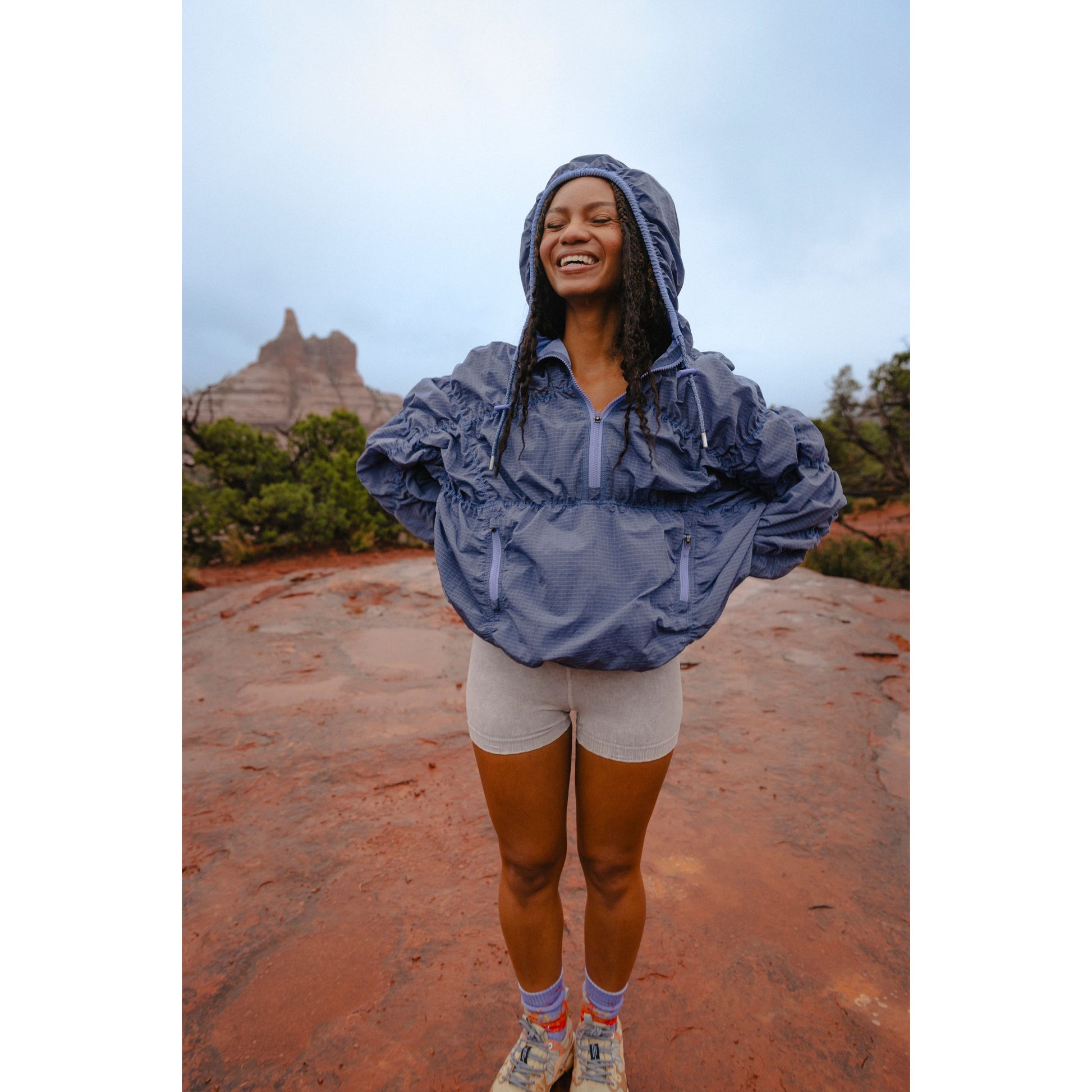 A joyful woman in a Happy Camper Pullover by Free People Movement, Twilight Violet color, stands on a red rocky terrain with a hood on her head, smiling under a cloudy sky.