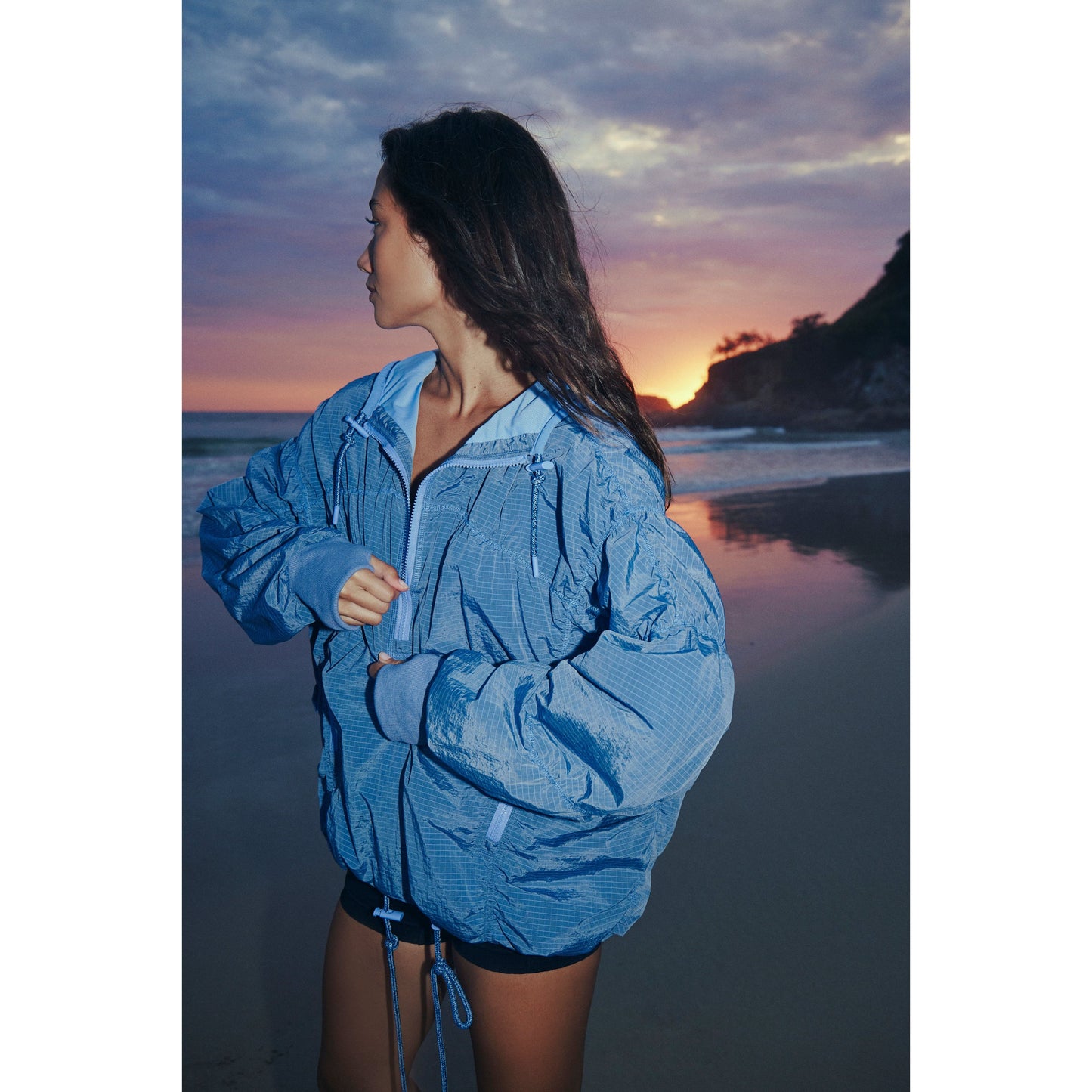 A woman in a wind- and water-resistant Happy Camper Pullover in Twilight Violet stands on a beach at sunset, looking over her shoulder toward the sky from Free People Movement.