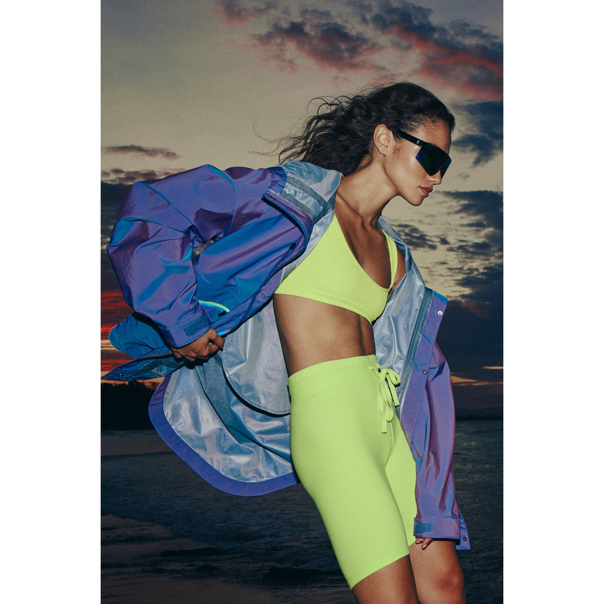A woman in sunglasses and colorful sportswear stands on a beach at sunset, with a dramatic sky in the background wearing the Singin in the Rain Jacket in Iridescent Blue Iris by Free People Movement.