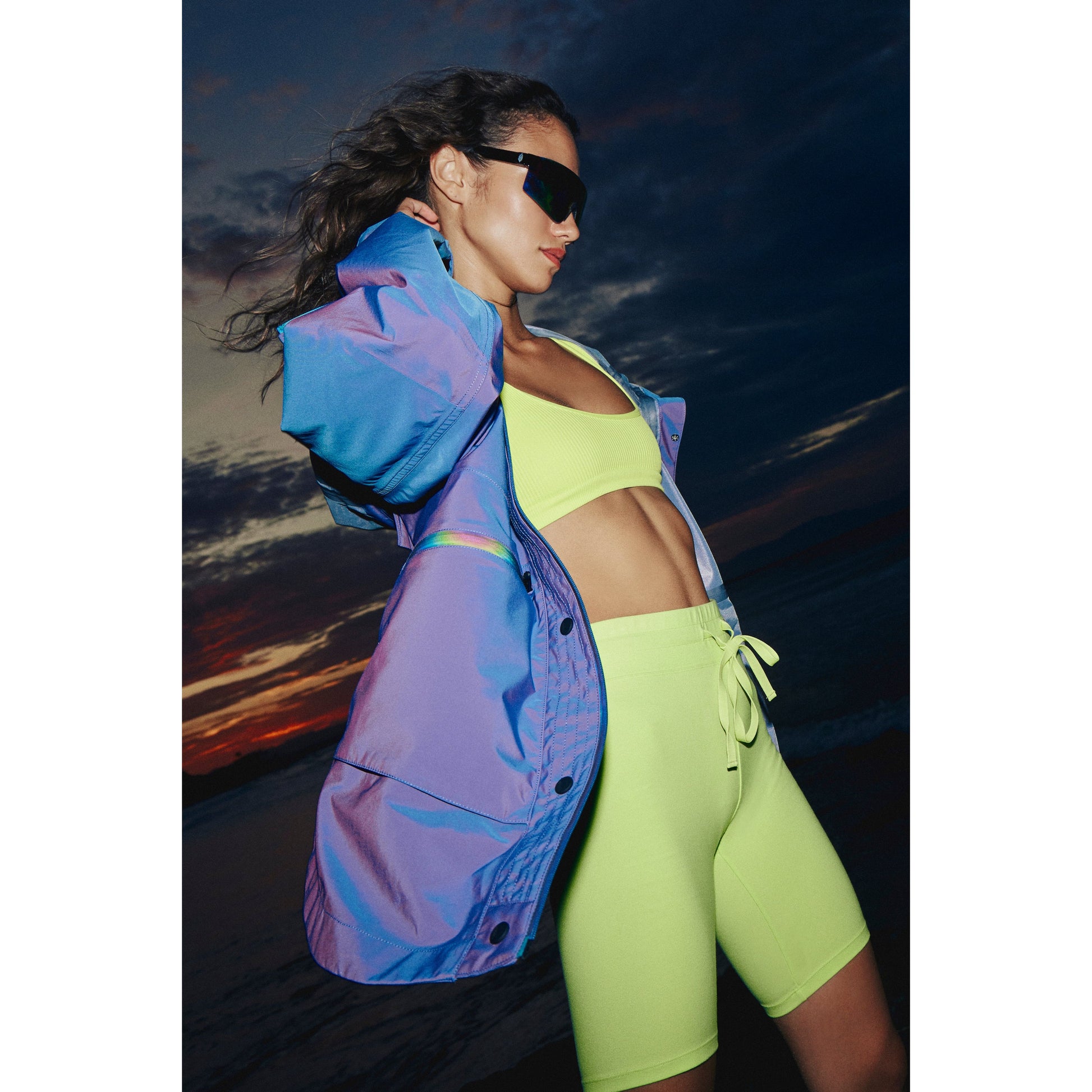 A woman in a Singin in the Rain Jacket, Iridescent Blue Iris by Free People Movement and sunglasses poses against a sunset sky.