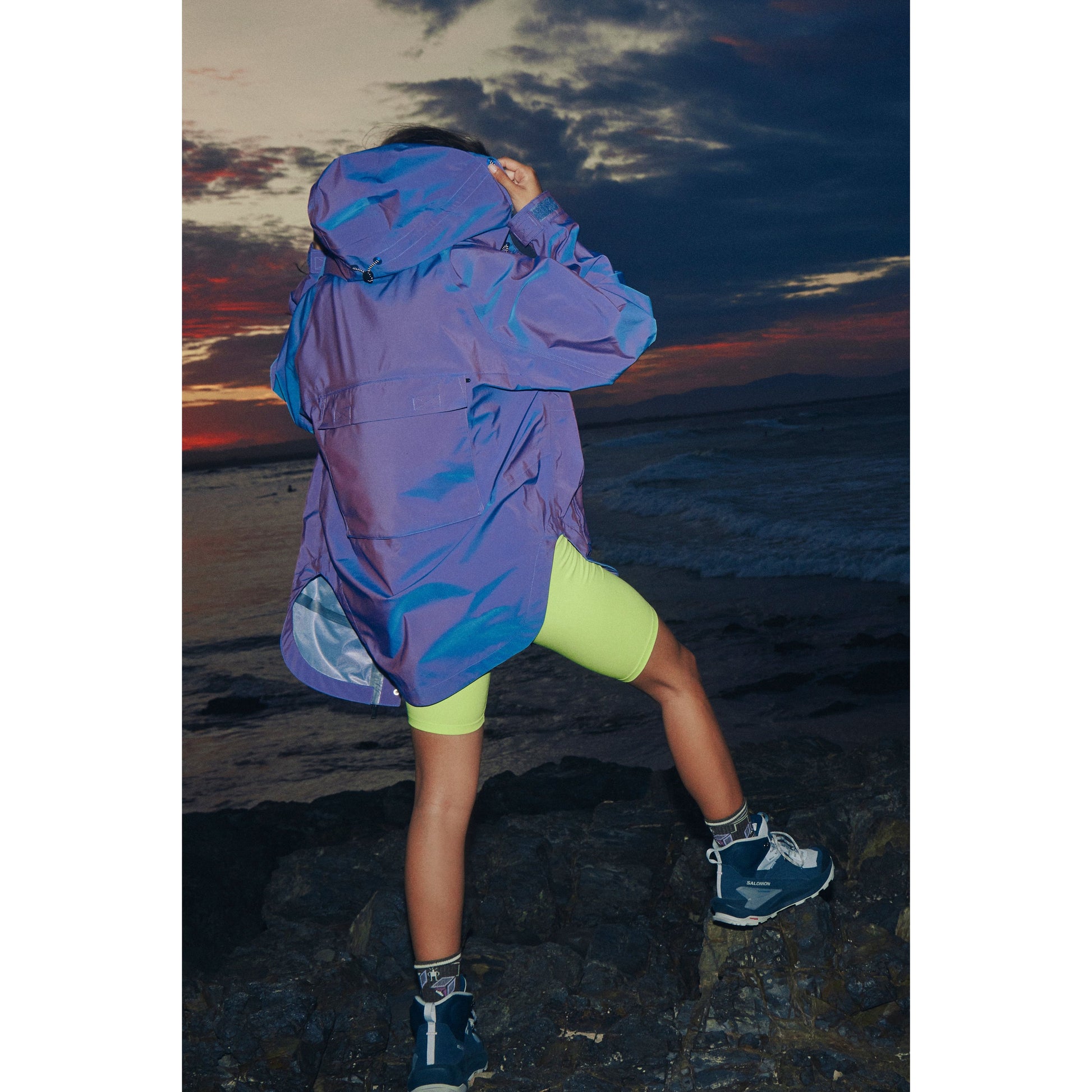 A person in a Singin in the Rain Jacket in Iridescent Blue Iris and yellow shorts poses on rocky terrain against a sunset sky.