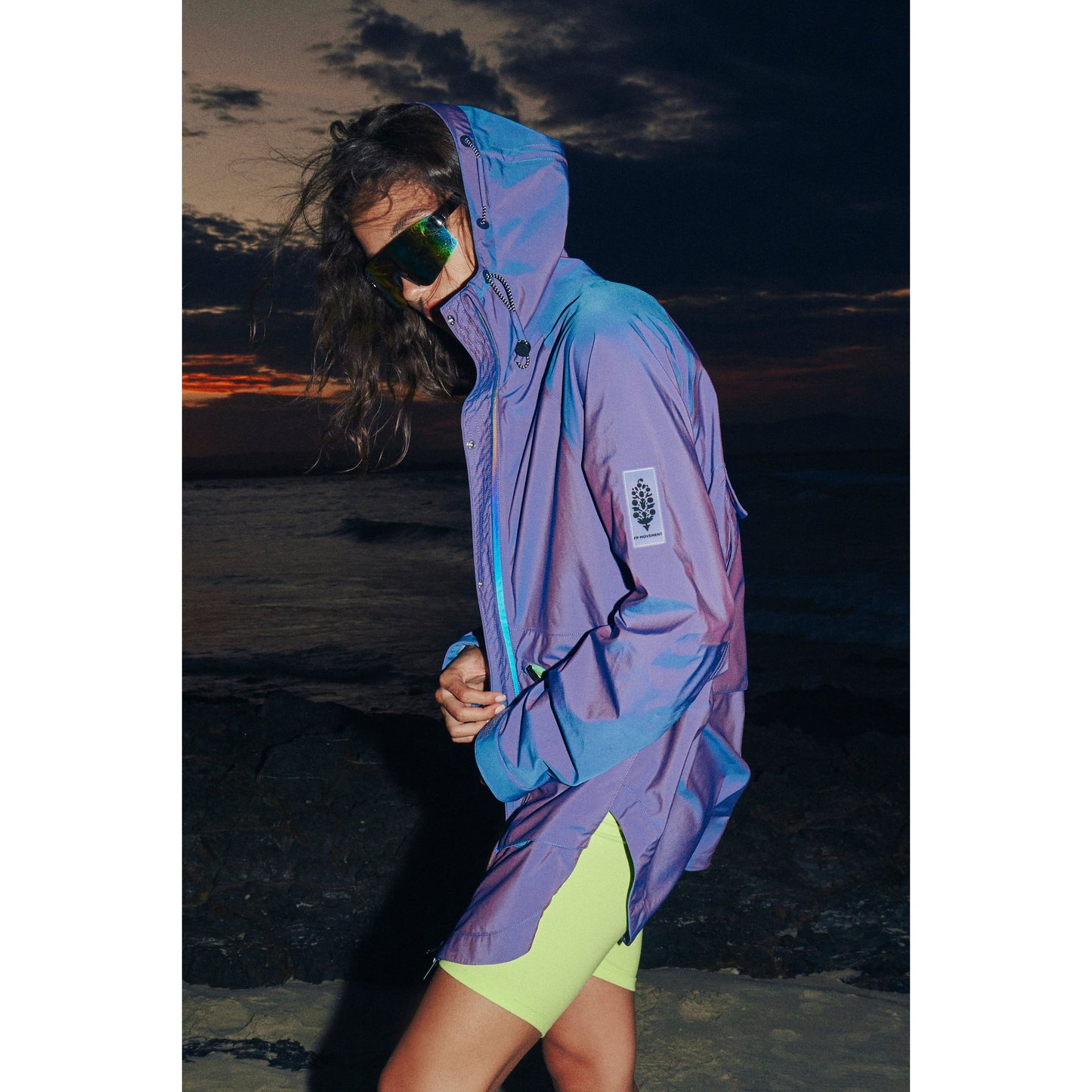 Woman in a Singin in the Rain Jacket, Iridescent Blue Iris by Free People Movement, and sunglasses standing by the sea at sunset, with her hair blowing in the wind.