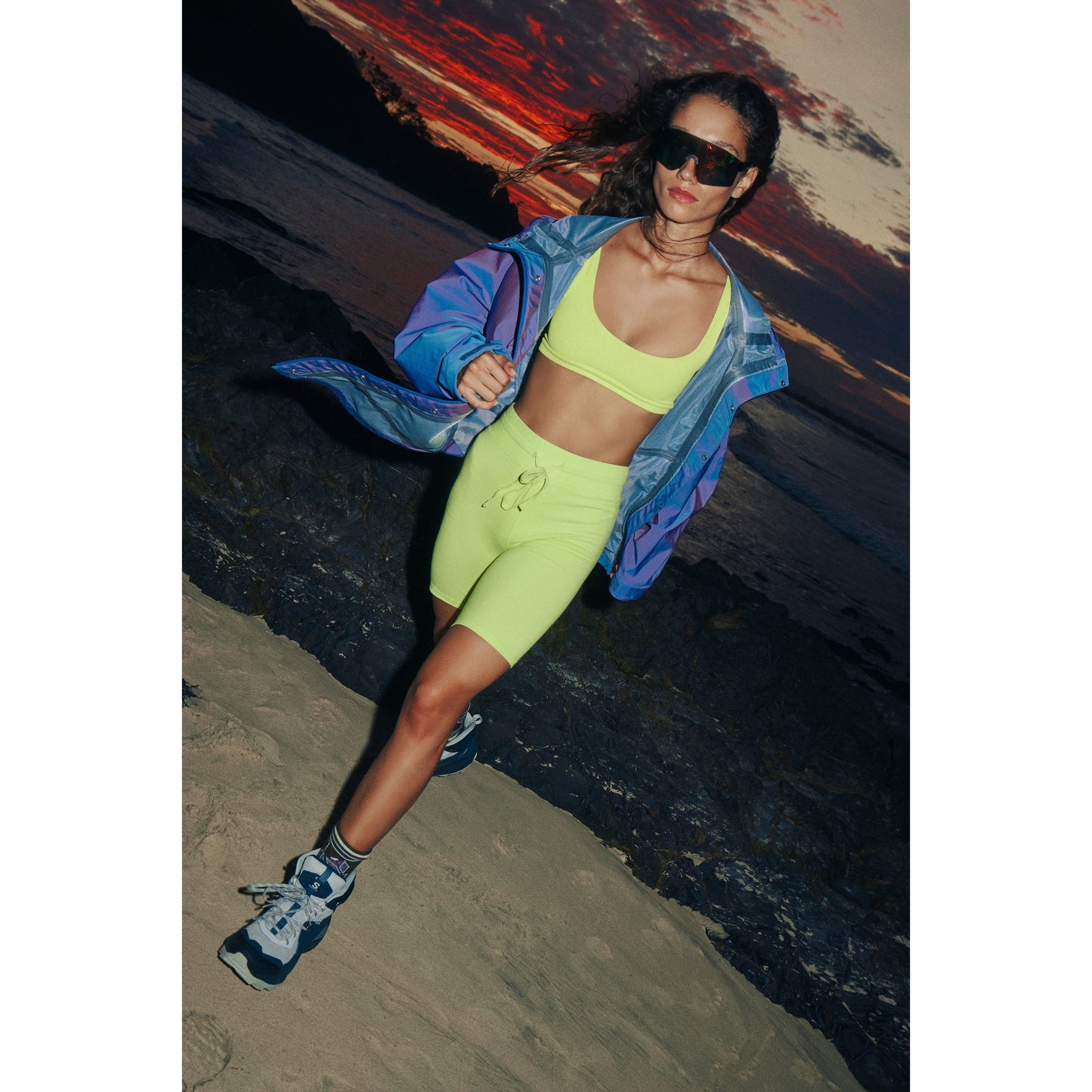 A woman in a lime green sports bra and shorts with a Singin in the Rain Jacket, Iridescent Blue Iris poses on a beach at sunset, wearing sunglasses from Free People Movement.